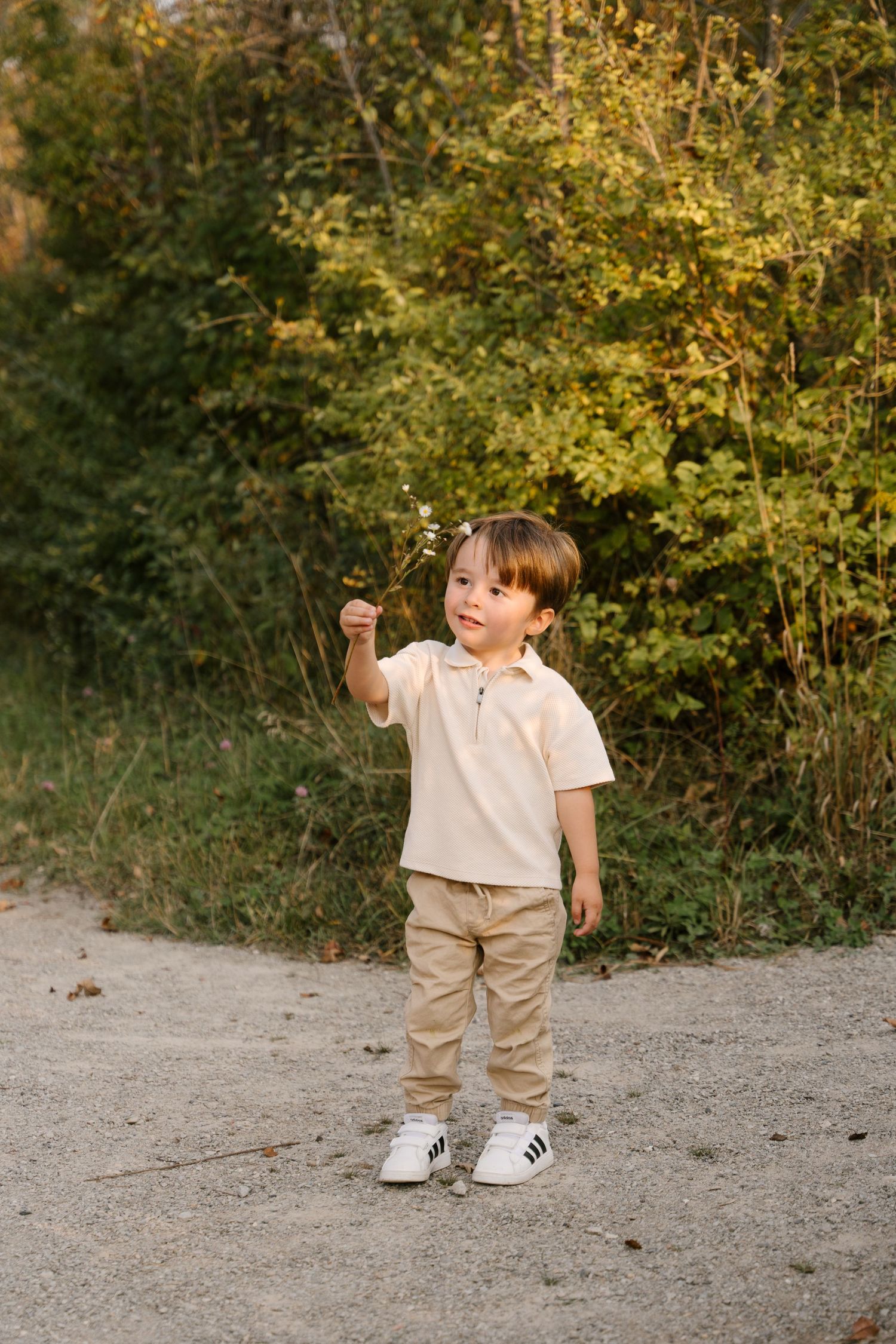 Cute toddler boy in beige outfit holds a small flower while standing on a path near green bushes.