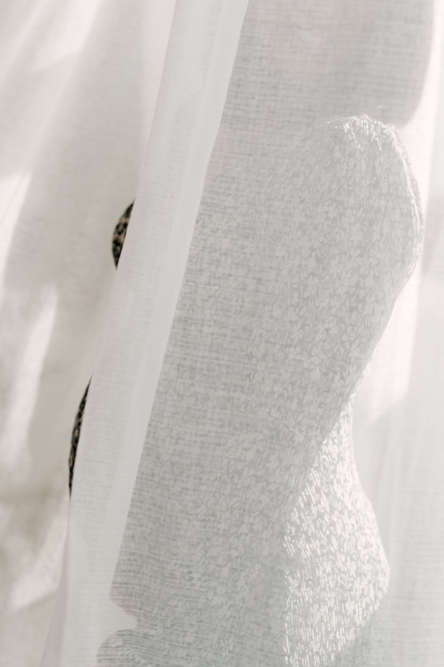 Silhouette of pregnant woman's baby bump visible through sheer white curtains in soft light.