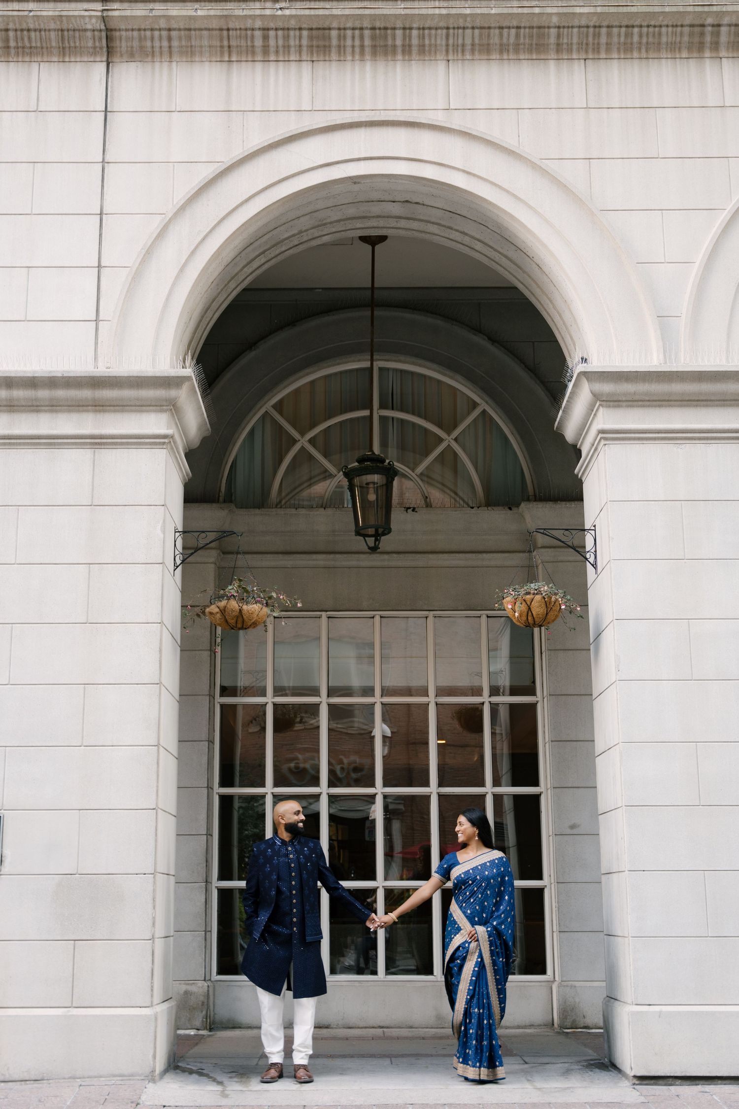 Couple in traditional Indian attire holding hands under an elegant arched entrance of a classic white building.
