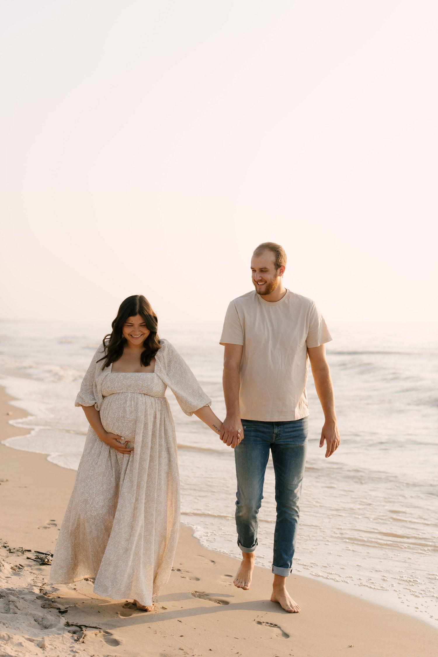 Pregnant couple holding hands walking along shoreline at golden hour sunset on sandy beach.