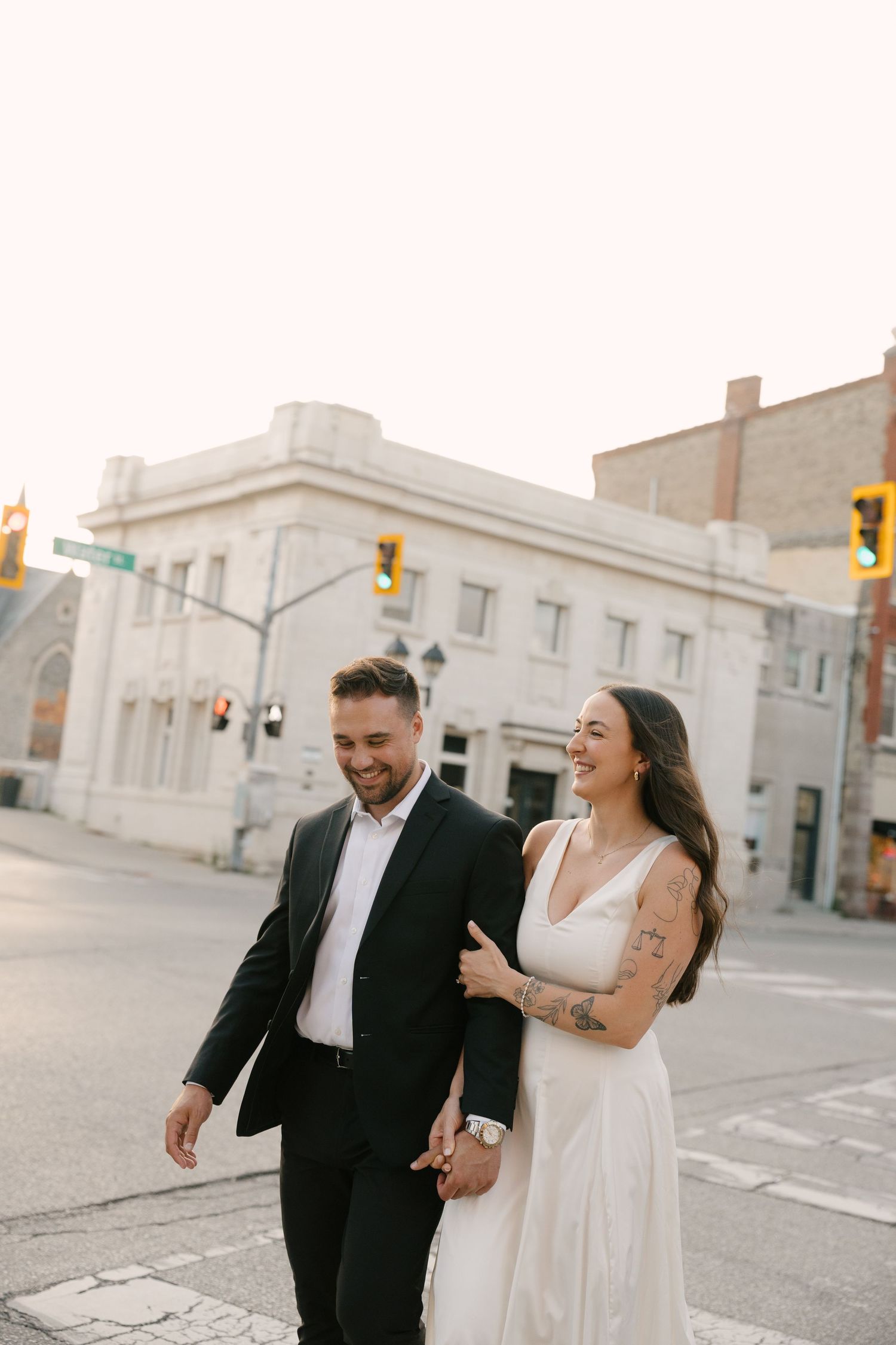 Smiling newlyweds walking hand in hand across urban intersection in wedding attire at golden hour.