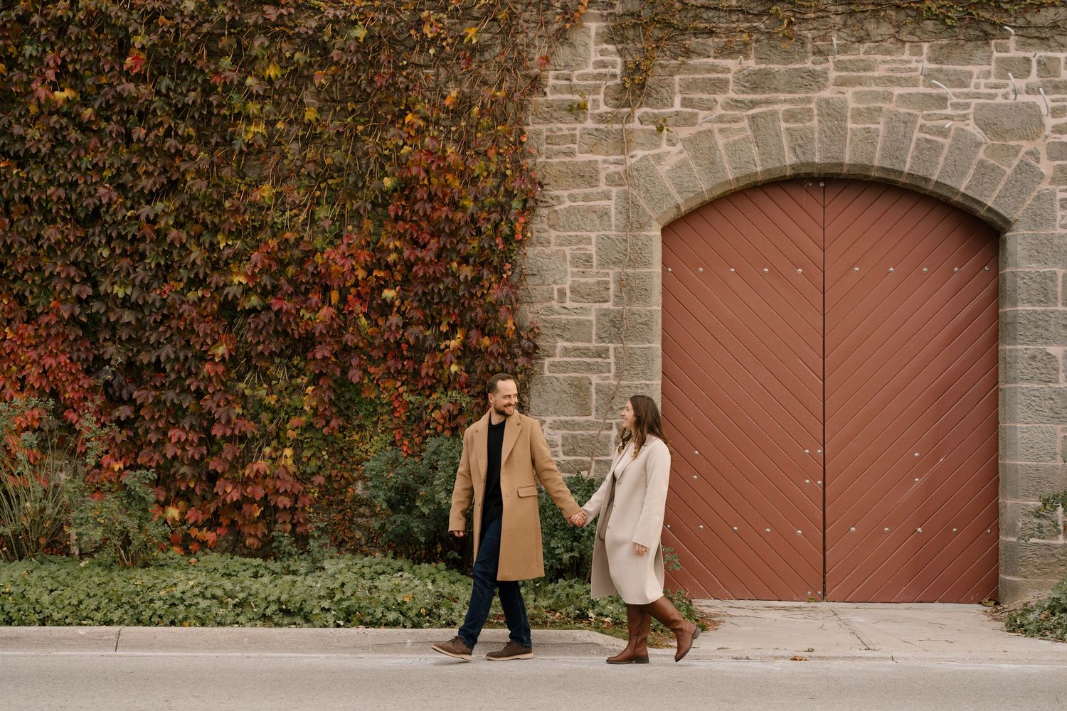 Couple holding hands walking past a stone building with colorful autumn ivy and red arched door.