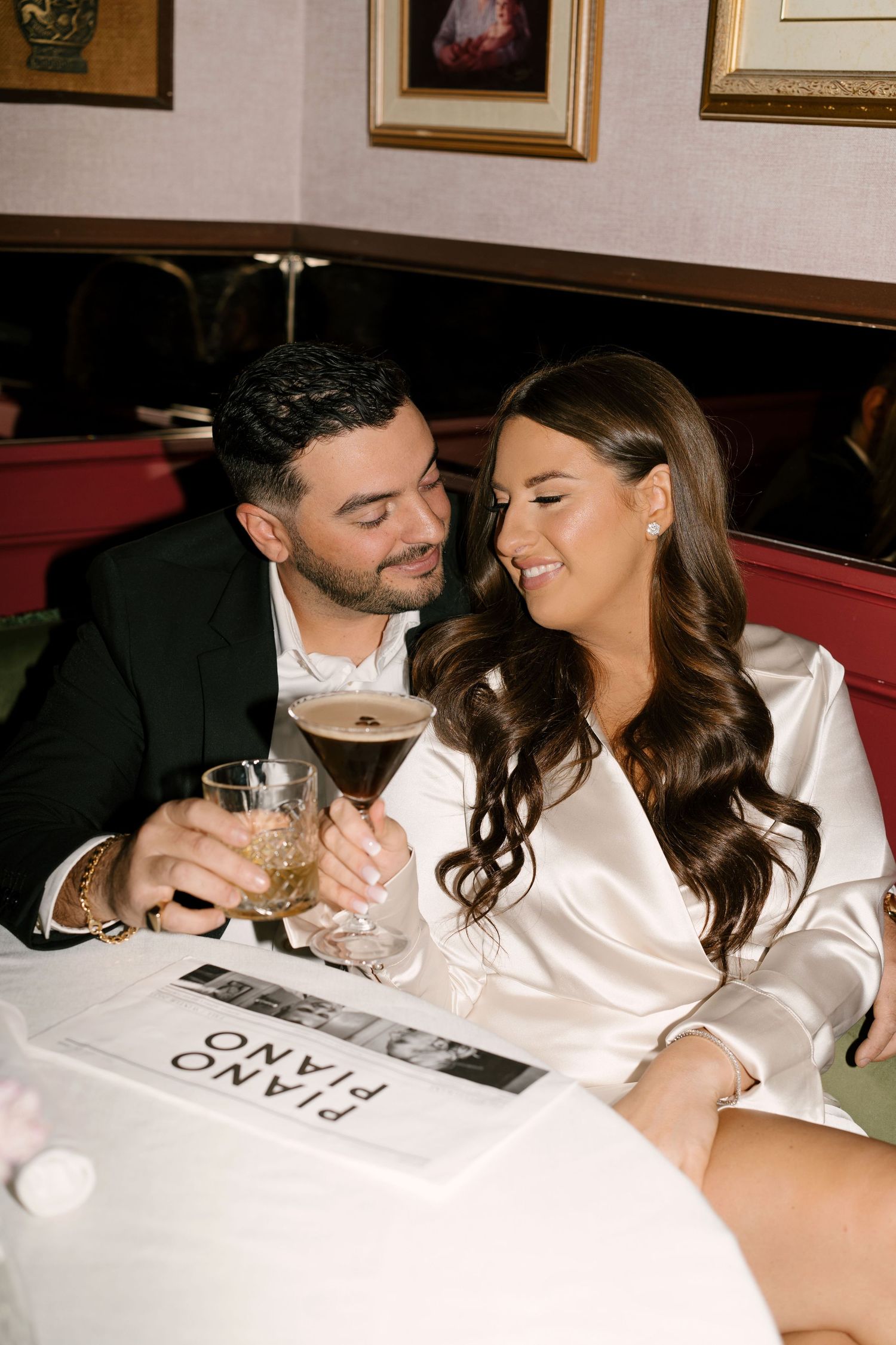 Couple toasting espresso martinis at Piano Piano restaurant booth, woman in white satin dress, man in black suit.