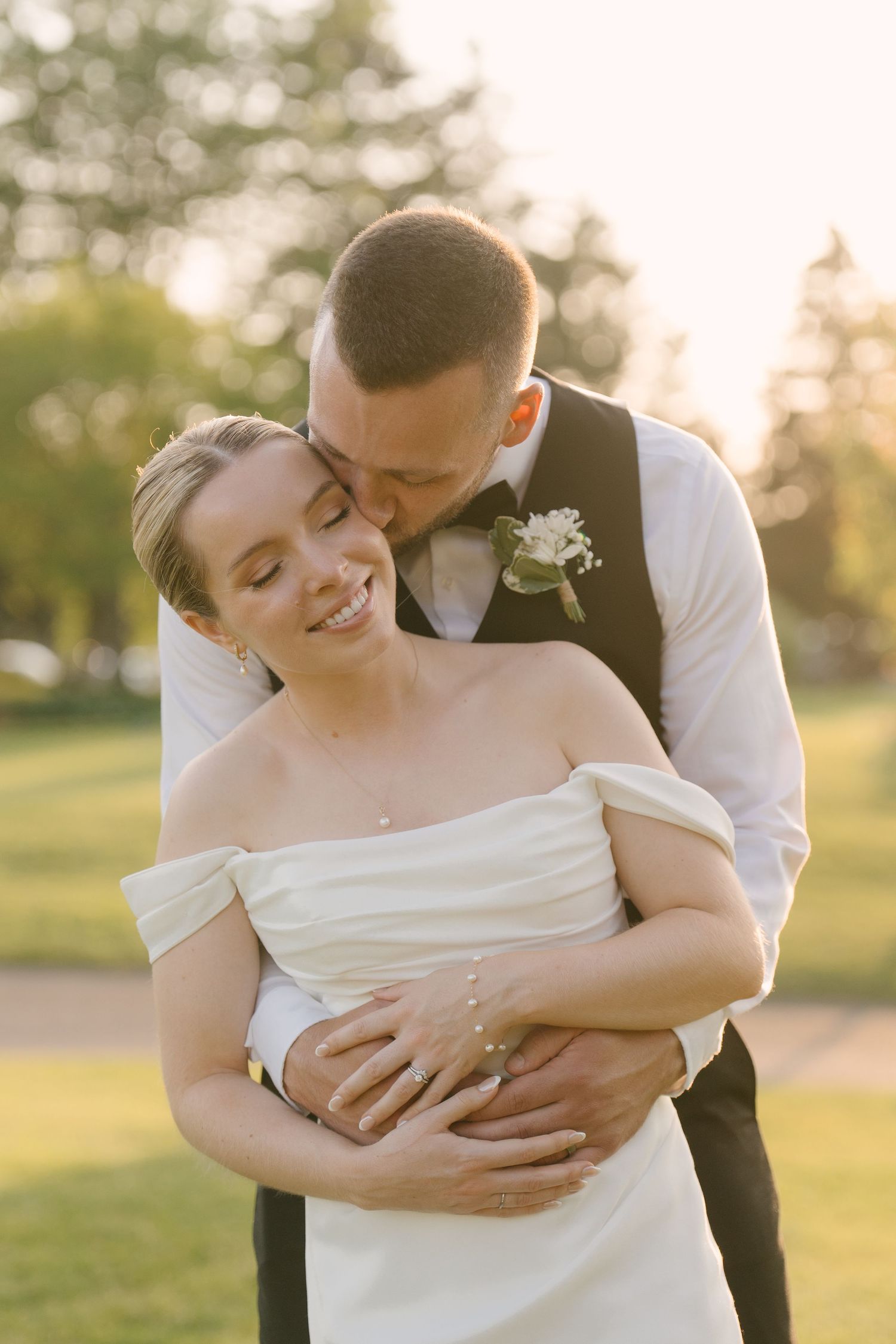 Groom embracing bride from behind in golden sunset light, bride wearing off-shoulder white gown, smiling joyfully outdoors.