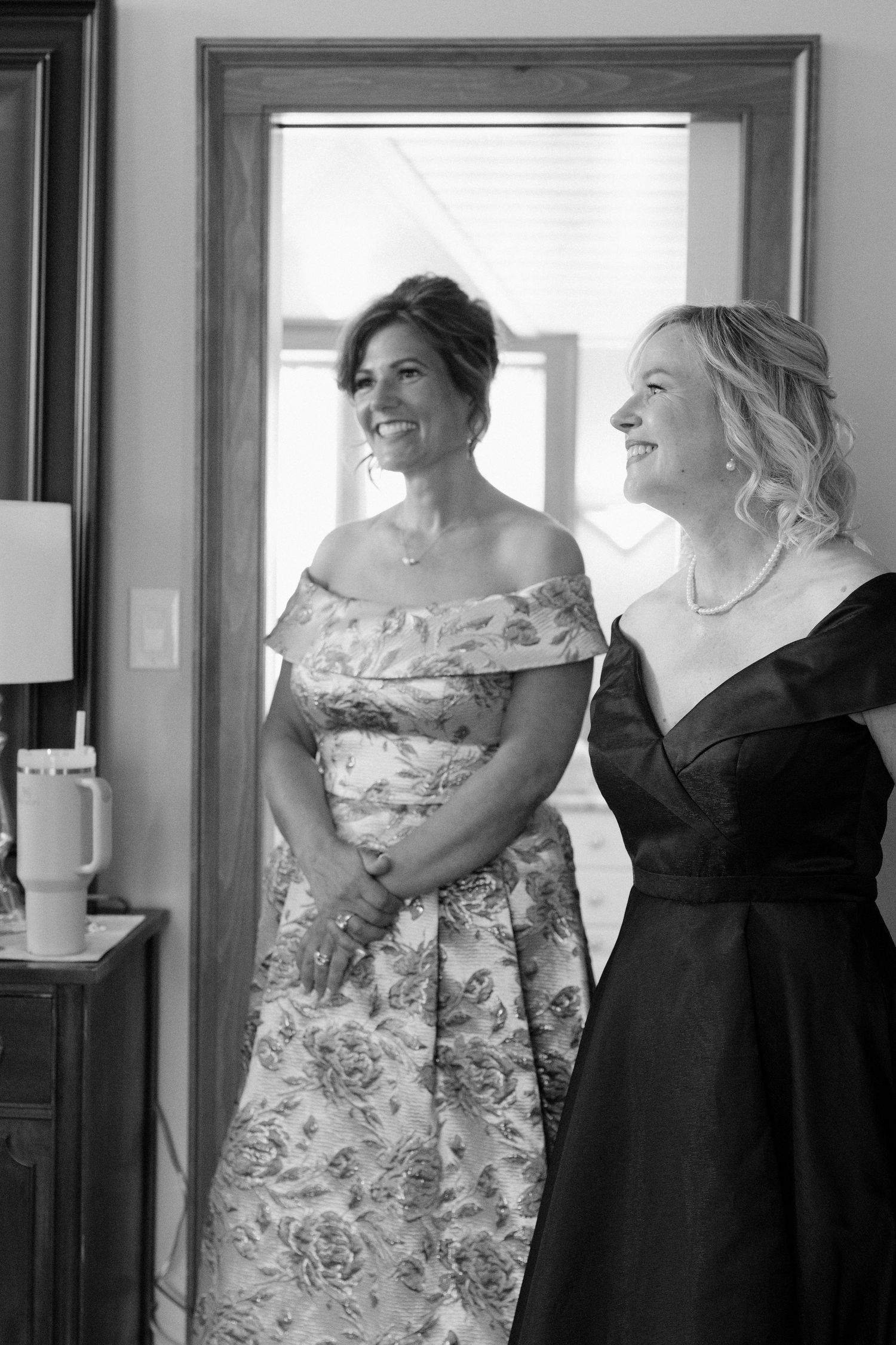 Two elegantly dressed women smile joyfully during wedding preparations indoors.