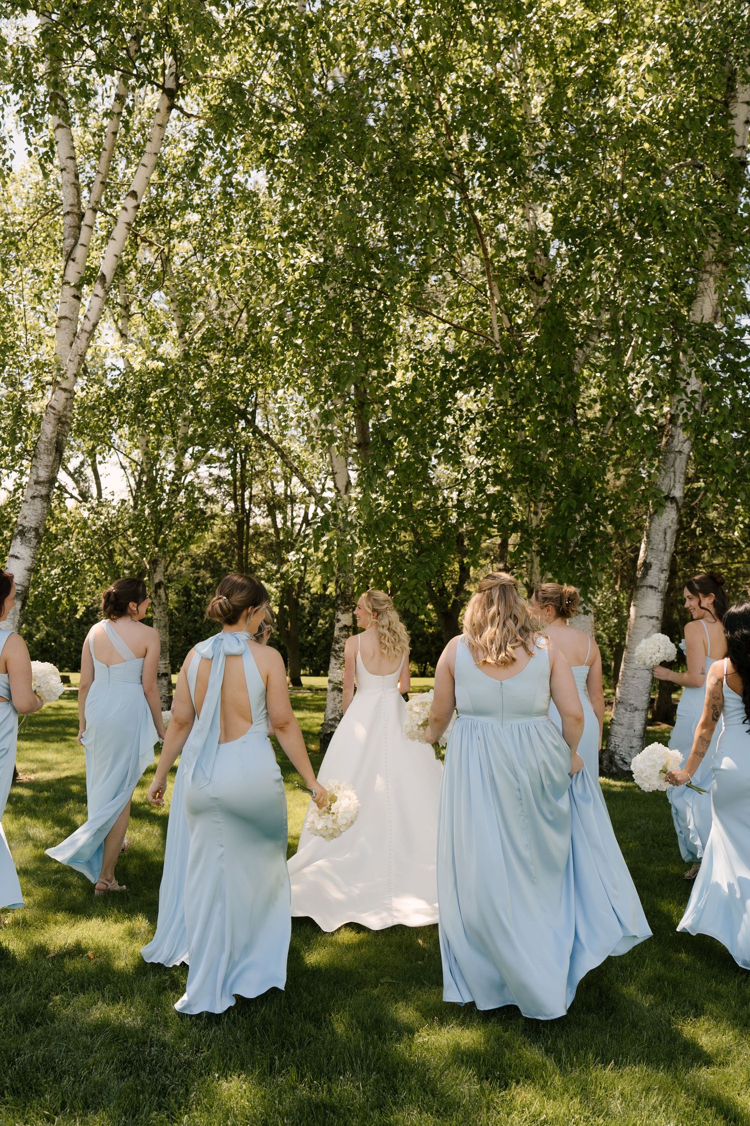 Bridesmaids in light blue gowns walk with bride in white dress through birch tree garden.