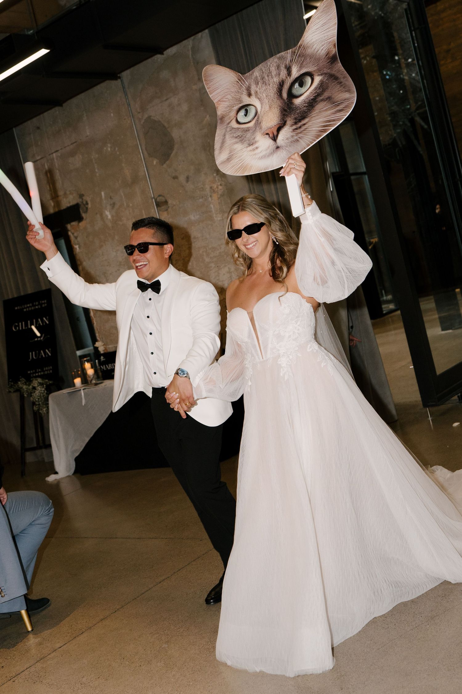 Laughing newlyweds in sunglasses make grand reception entrance holding giant cat face fan and sparklers at Tapestry Hall wedding venue.