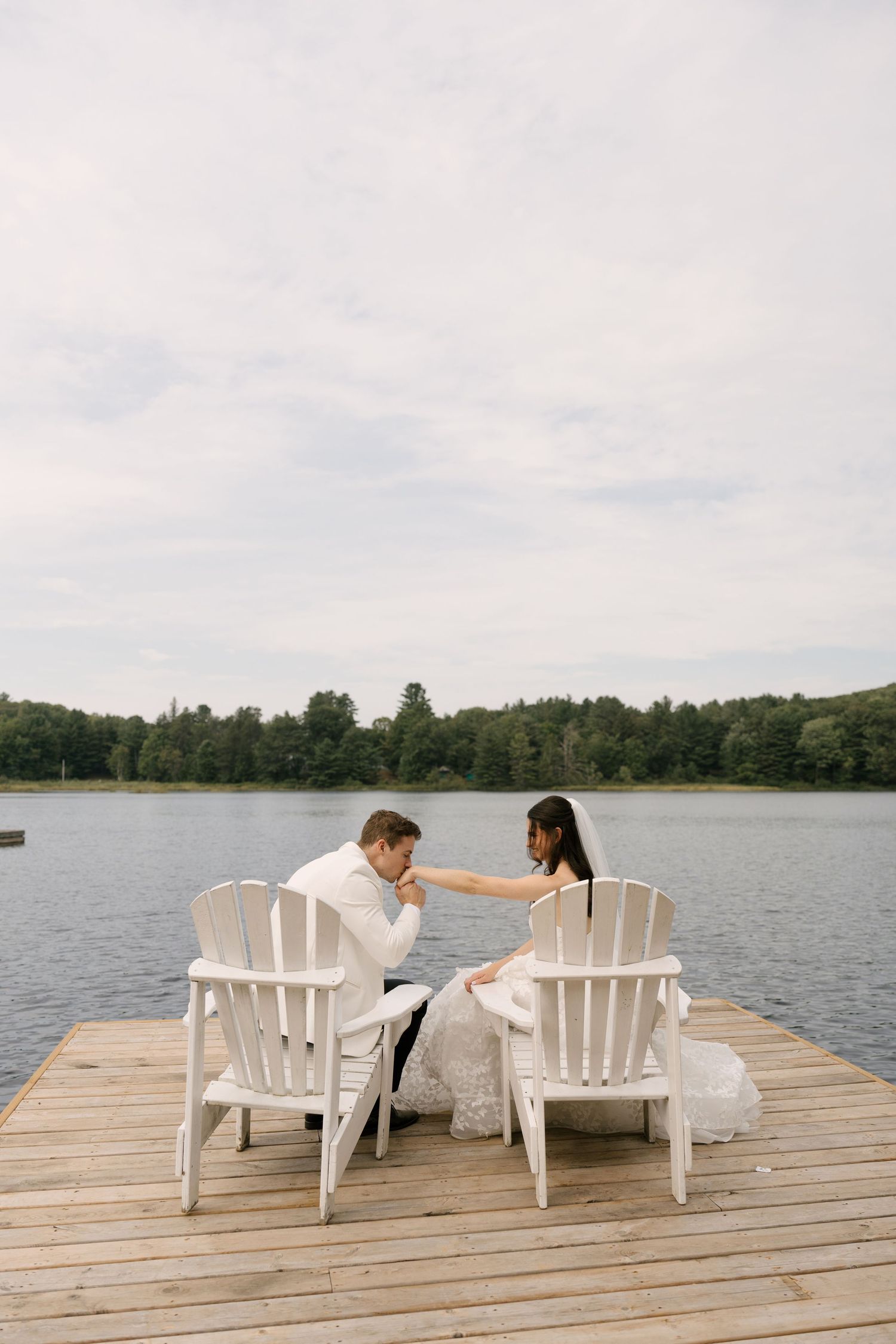 Bride and groom sit in white Adirondack chairs on a lakeside dock, sharing a tender moment on their wedding day.