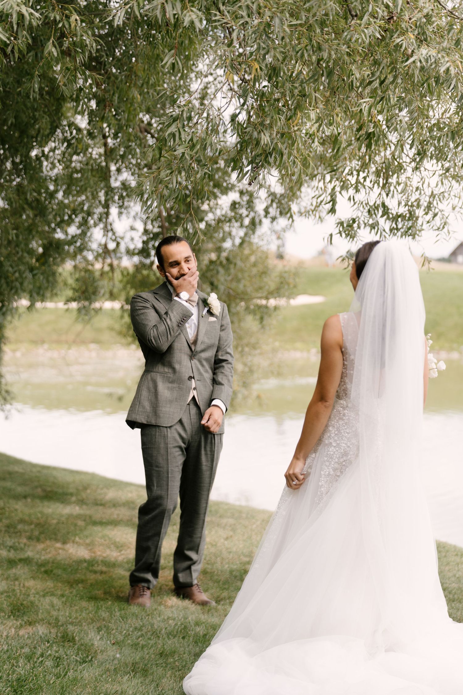 Groom in gray suit reacts emotionally seeing bride in white lace gown and veil during first look outdoors.
