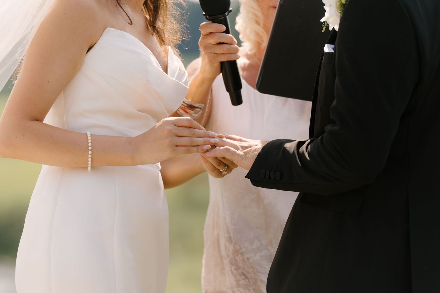 Bride places wedding ring on grooms hand during outdoor ring exchange ceremony, officiant holding microphone nearby.