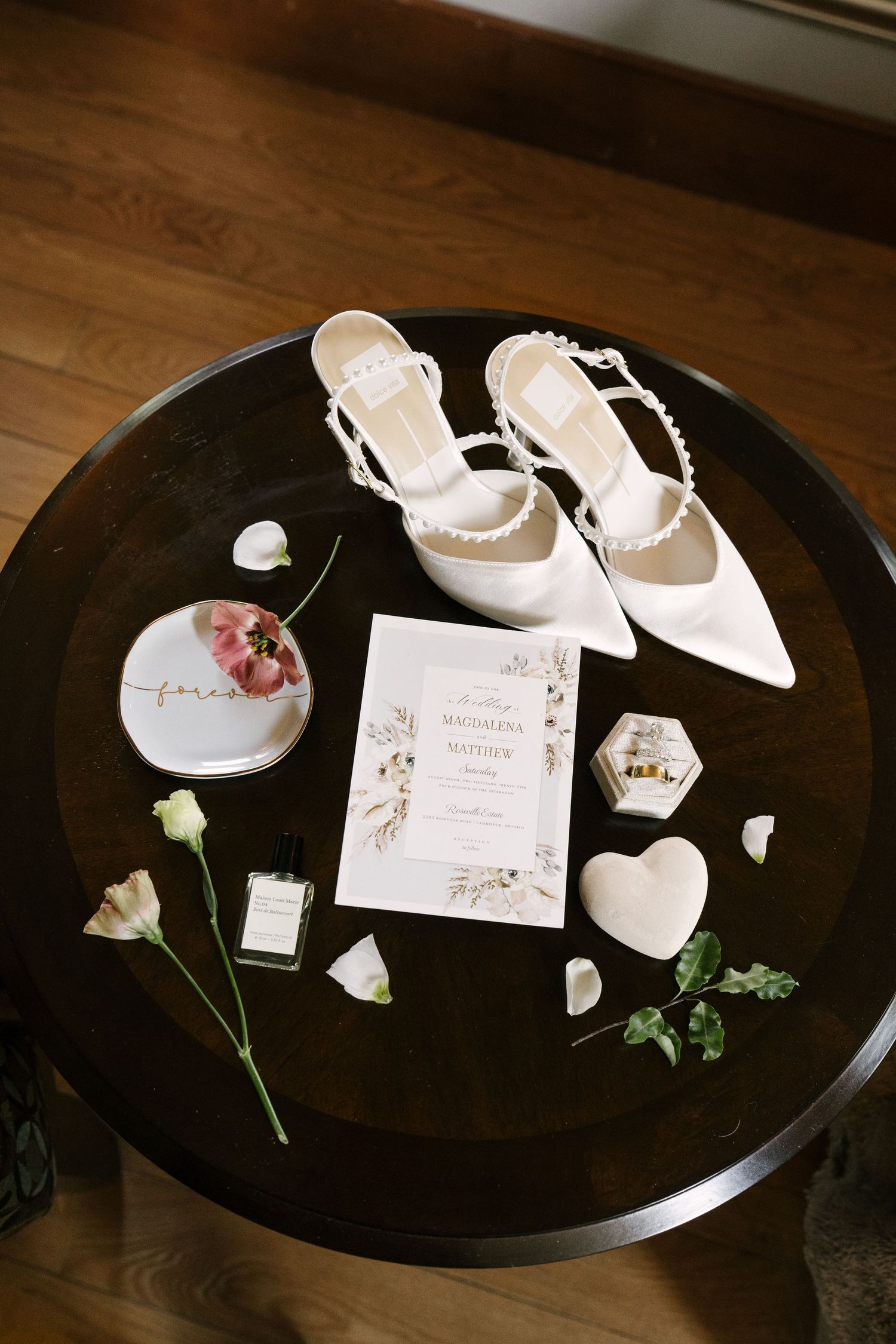 Flat lay of bridal accessories including white heels, invitation, and rings on table.