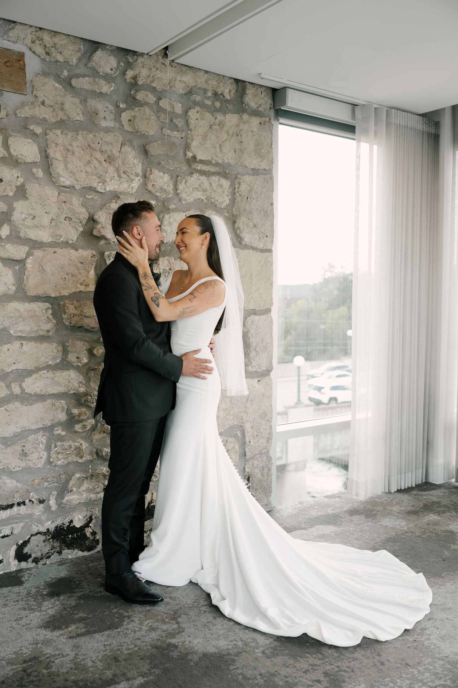Bride in white gown and veil embraces groom in black suit against stone wall near window at Cambridge Mill.