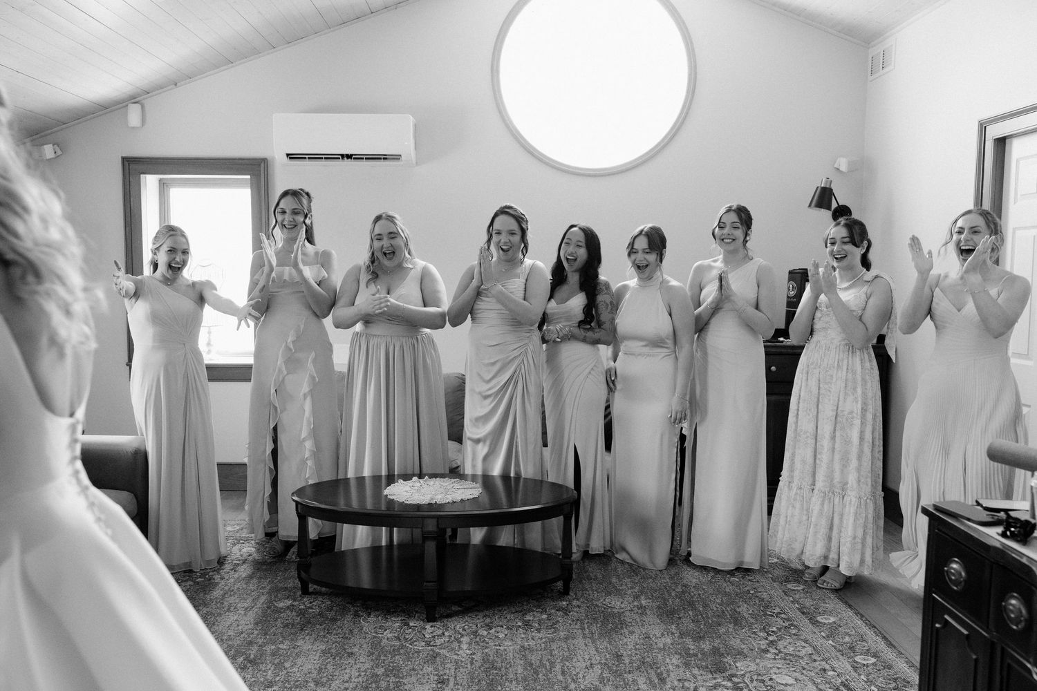 Bridesmaids in mismatched gowns reacting with excitement to bride's first look in black and white photo.
