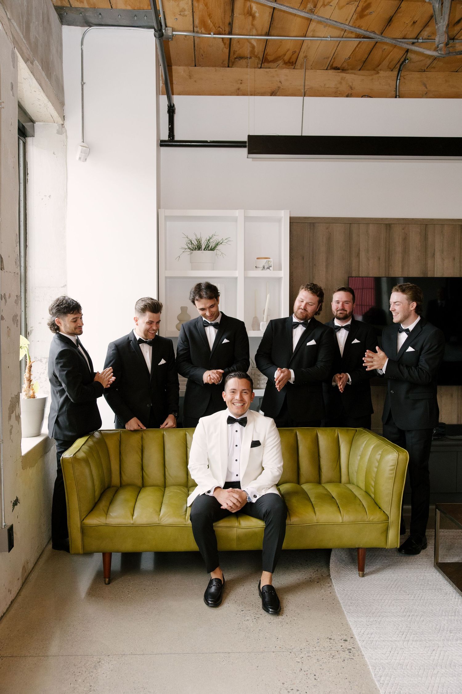 Groom in white tuxedo sits on green velvet sofa surrounded by groomsmen in black tuxedos laughing at Tapestry Hall wedding venue.