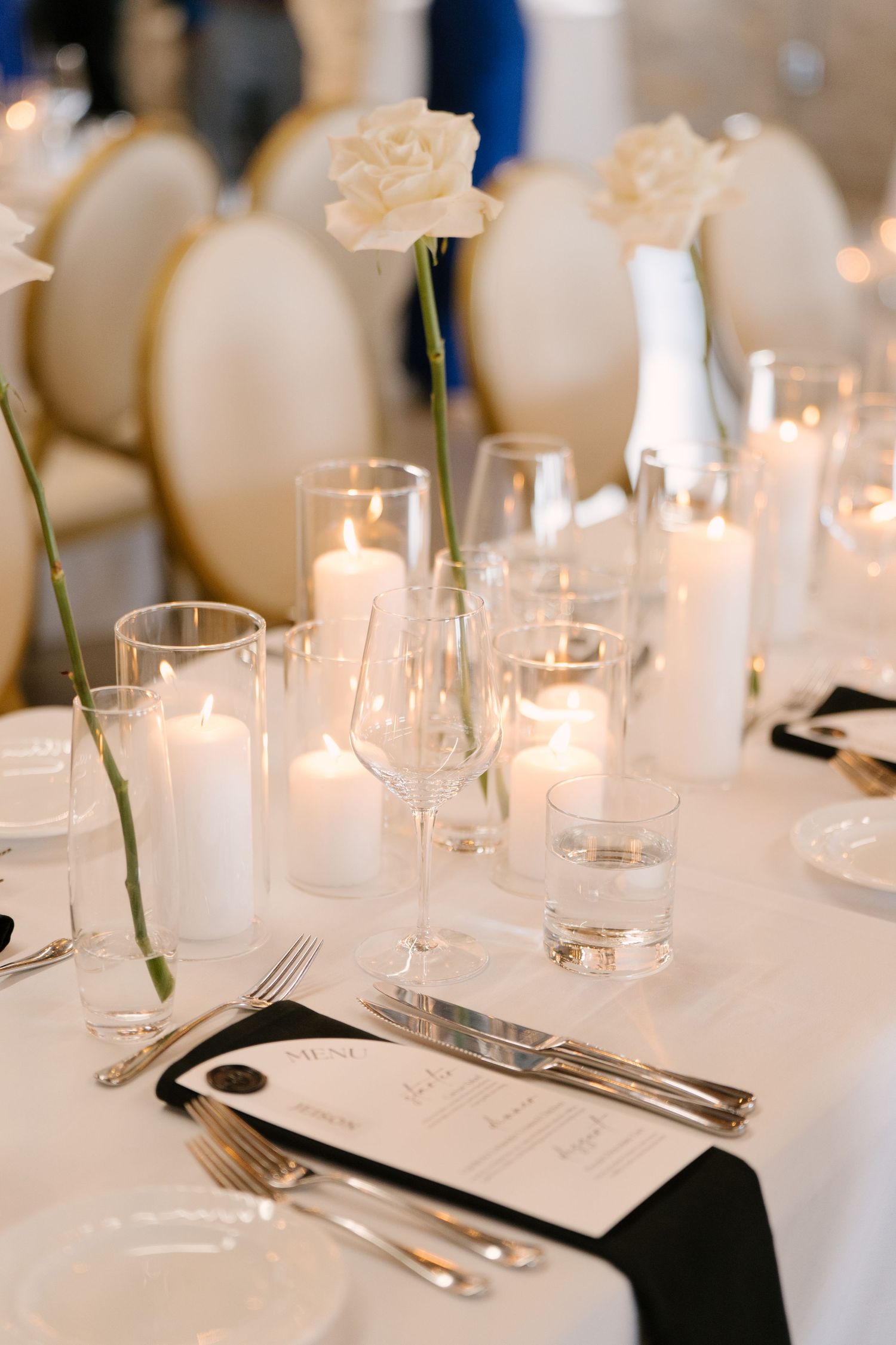 Romantic wedding table setting with white pillar candles, crystal glasses, black napkins, and single white rose centerpiece.