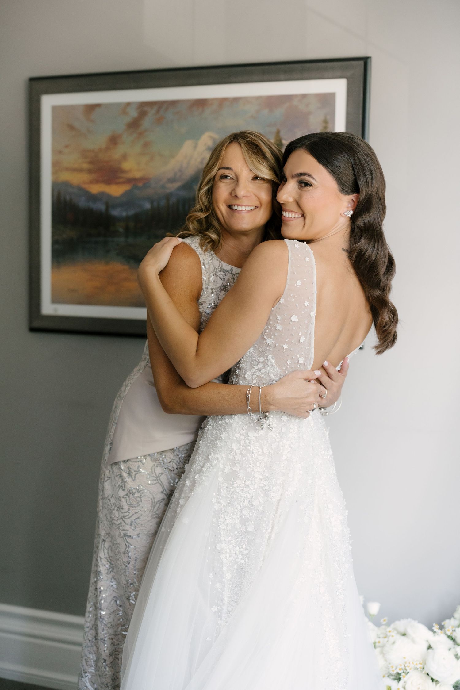 Bride in backless gown embraces smiling woman in silver dress before wedding ceremony.