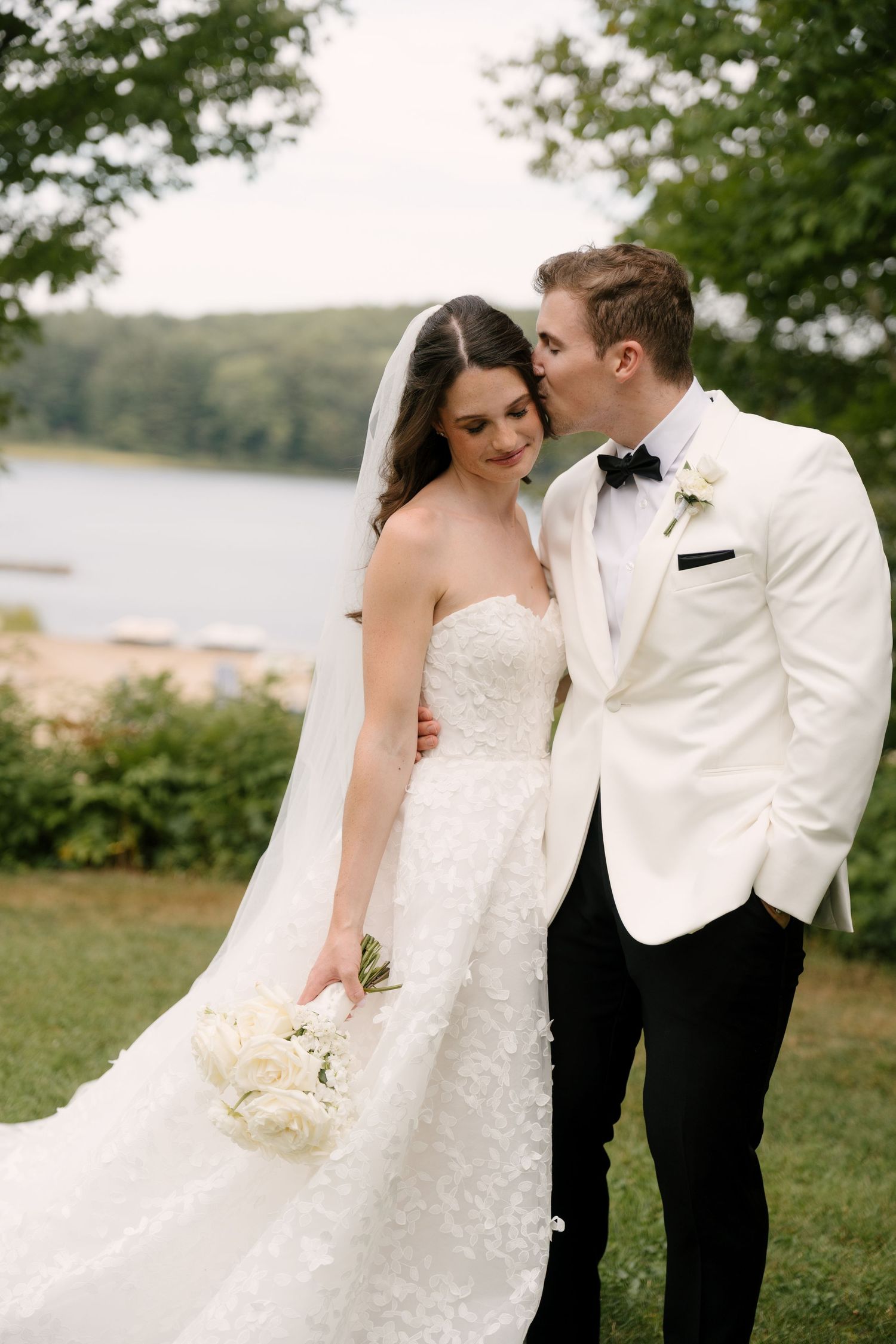 Groom in white tuxedo kisses bride wearing strapless lace gown holding white bouquet outdoors near lake.