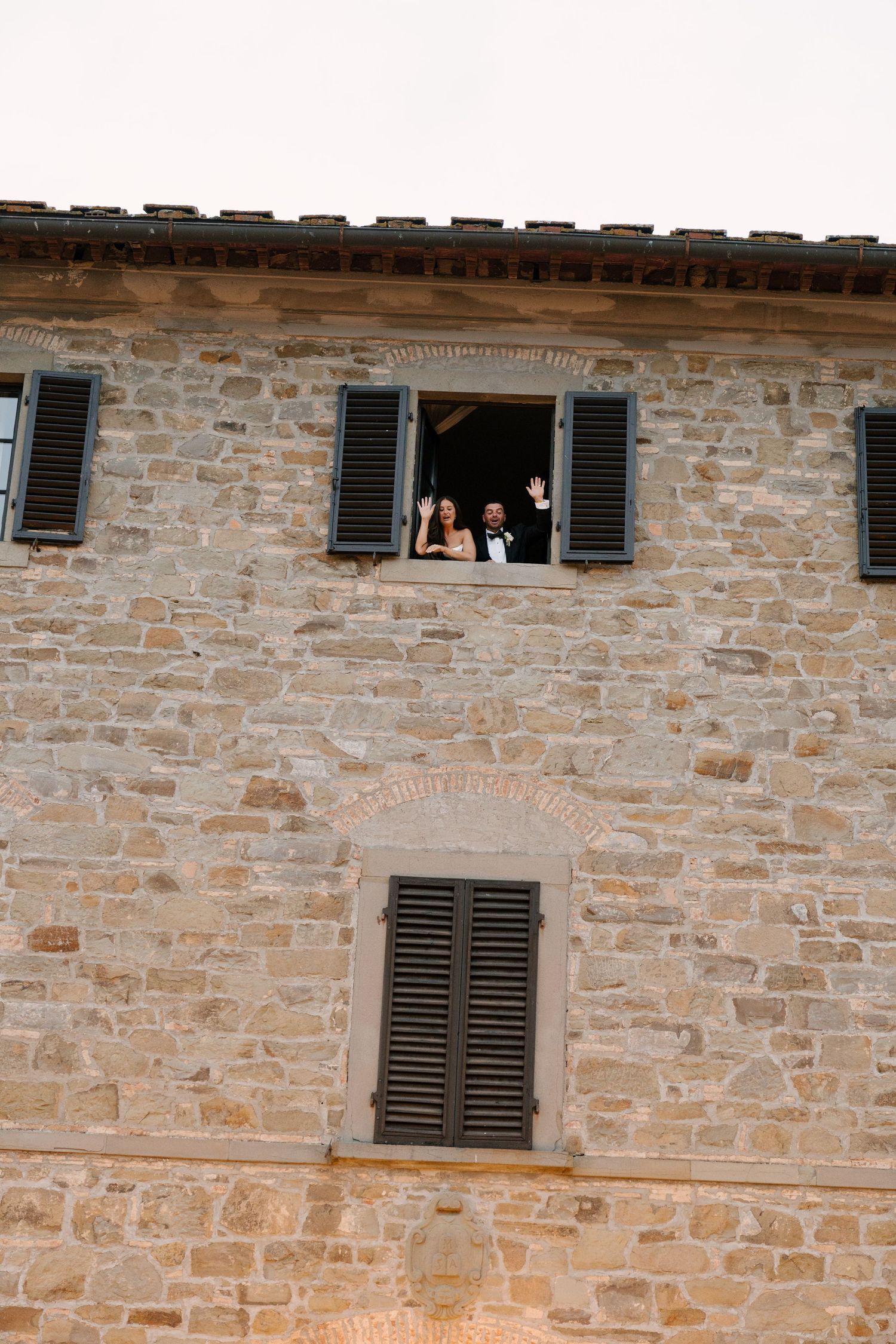 Bride and groom waving from open window of rustic stone Tuscan villa wedding venue.