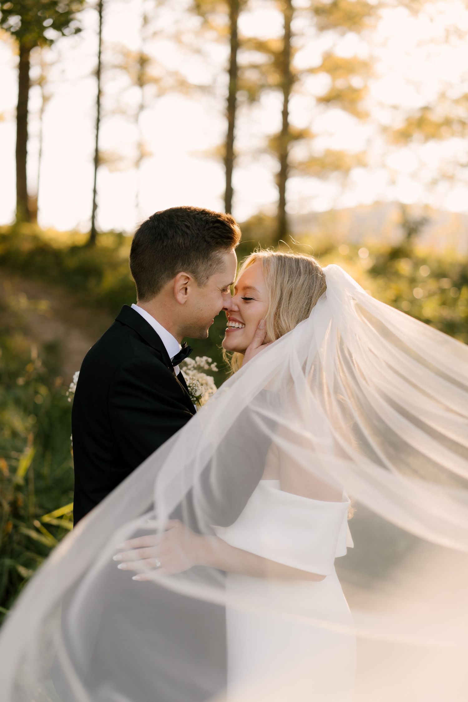 Bride and groom share an intimate moment as her flowing veil billows around them in golden sunset light.