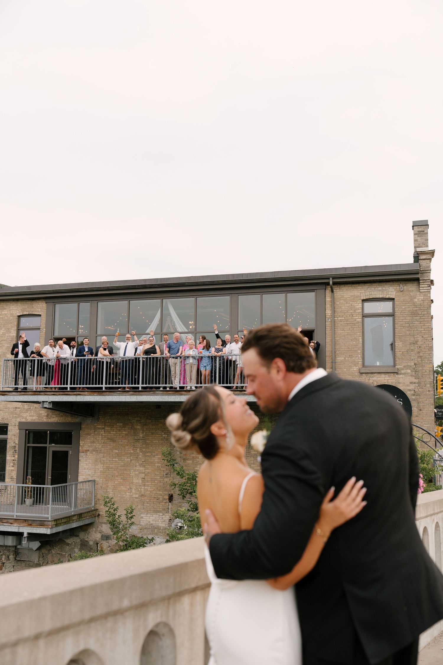 Bride and groom share first kiss while wedding guests watch from balcony above.