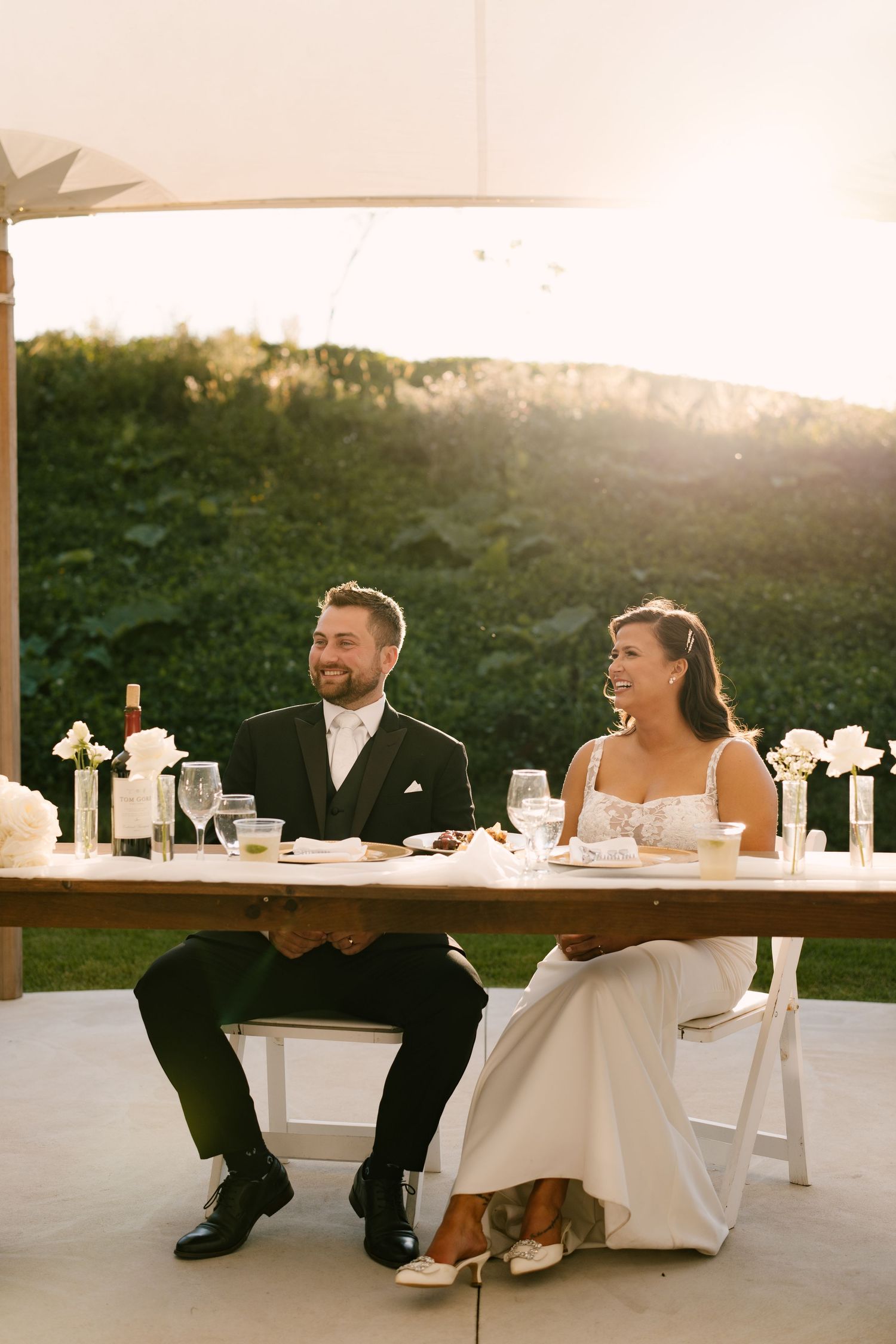 Bride and groom laughing together at sweetheart table during outdoor wedding reception at golden hour. Wedding venue at Wakefield Estates.