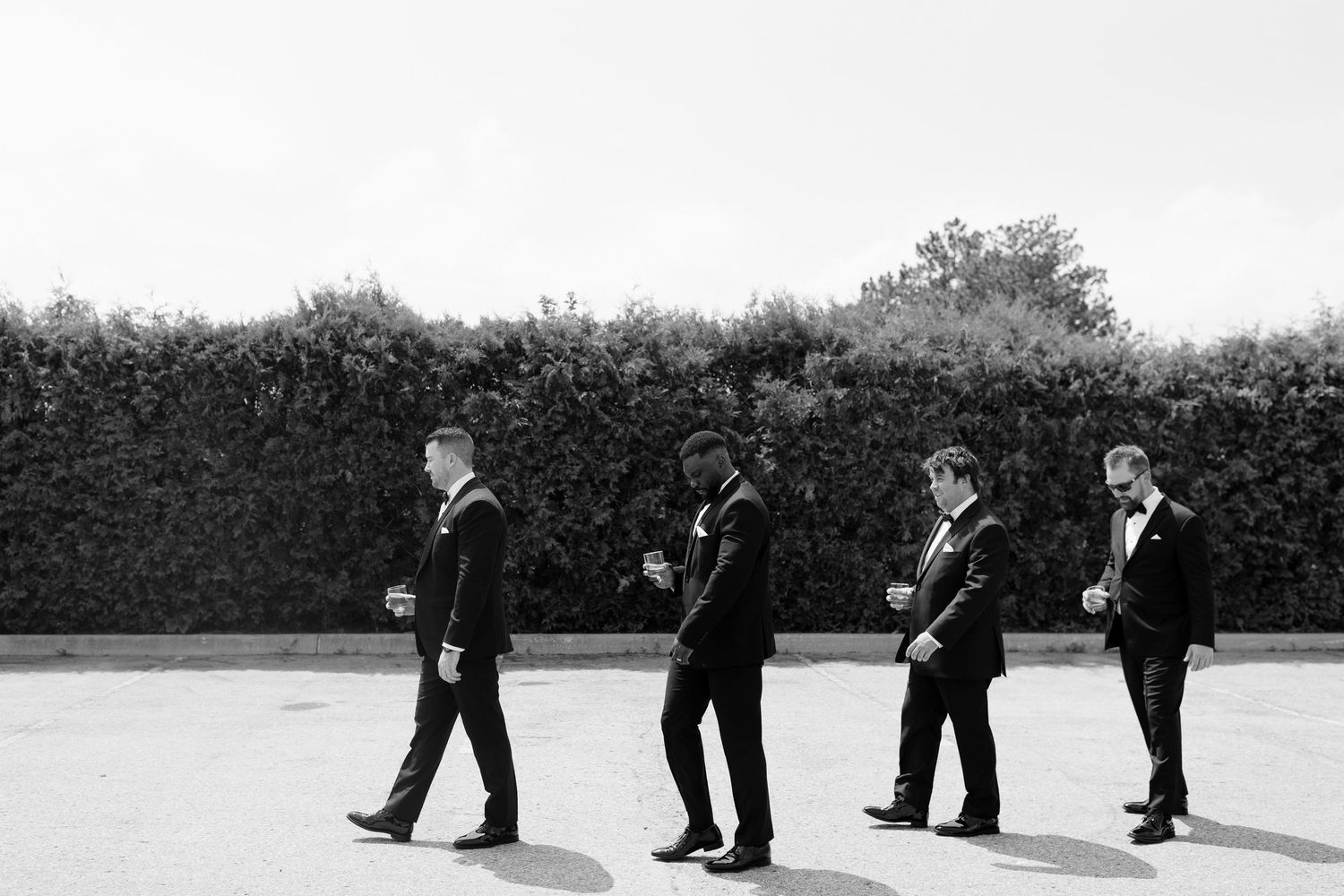 Black and white photo of groom and groomsmen in tuxedos walking confidently in a line outdoors.