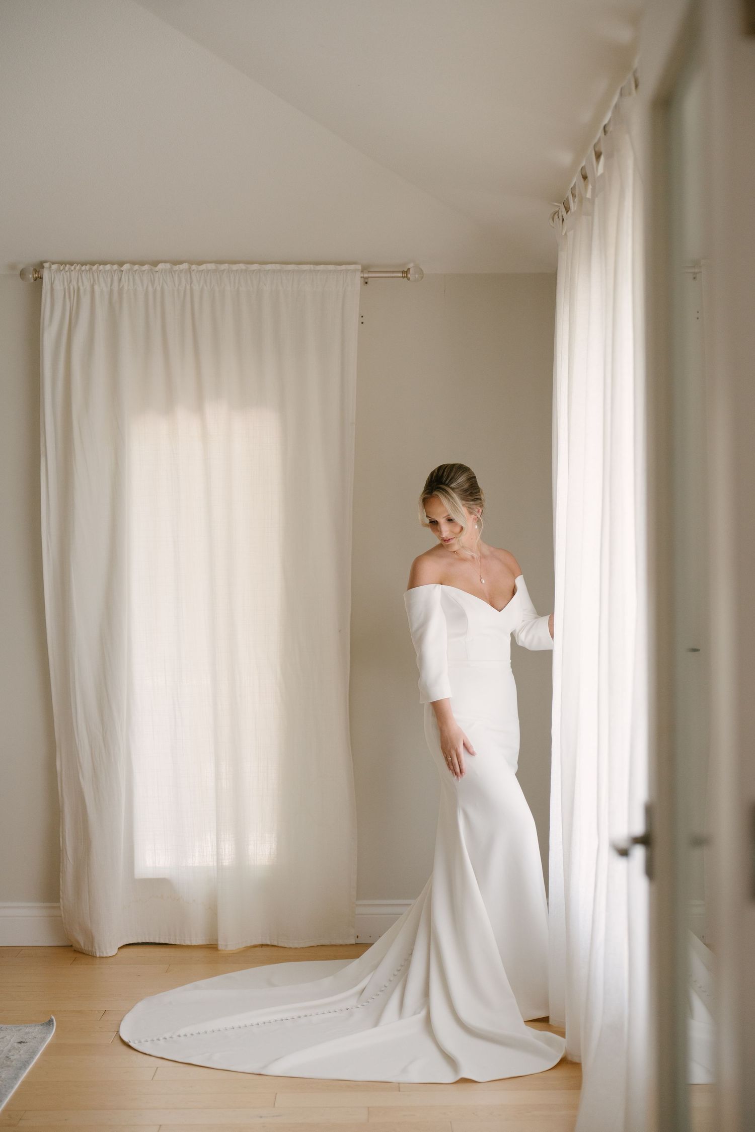 Bride in a fitted off-shoulder white wedding gown with a train standing by sheer curtains in a bright room.
