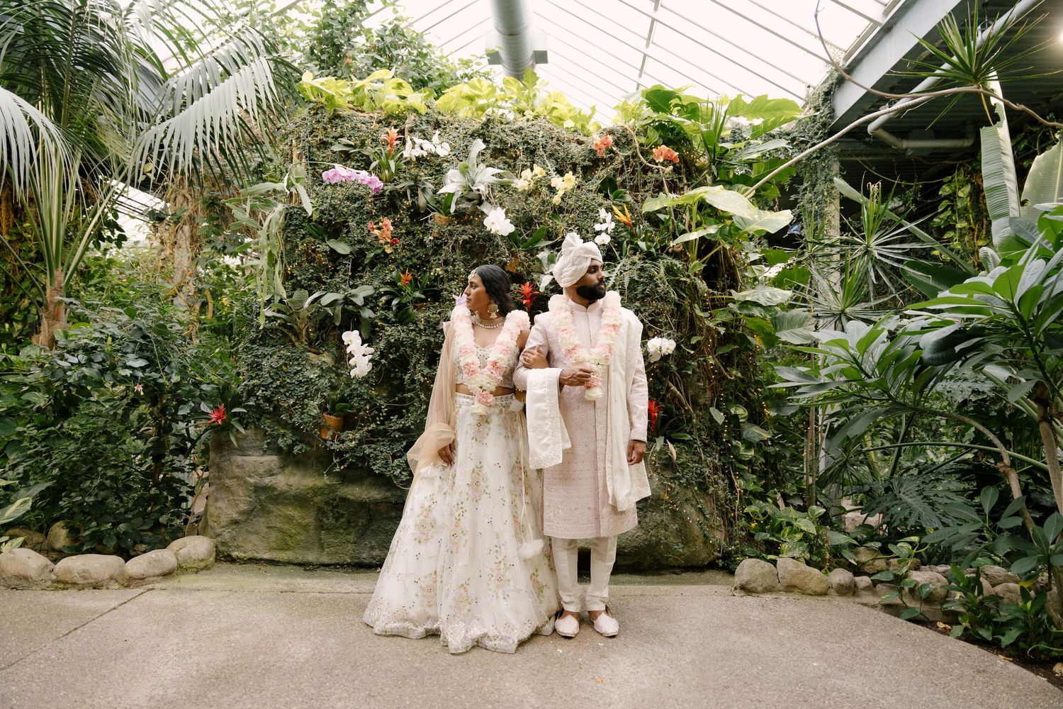 South Asian bride and groom in traditional wedding outfits stand together beneath a floral arch in a butterfly botanical garden.