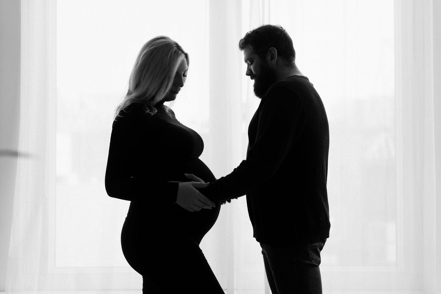Black and white silhouette of pregnant couple holding baby bump together near bright window.