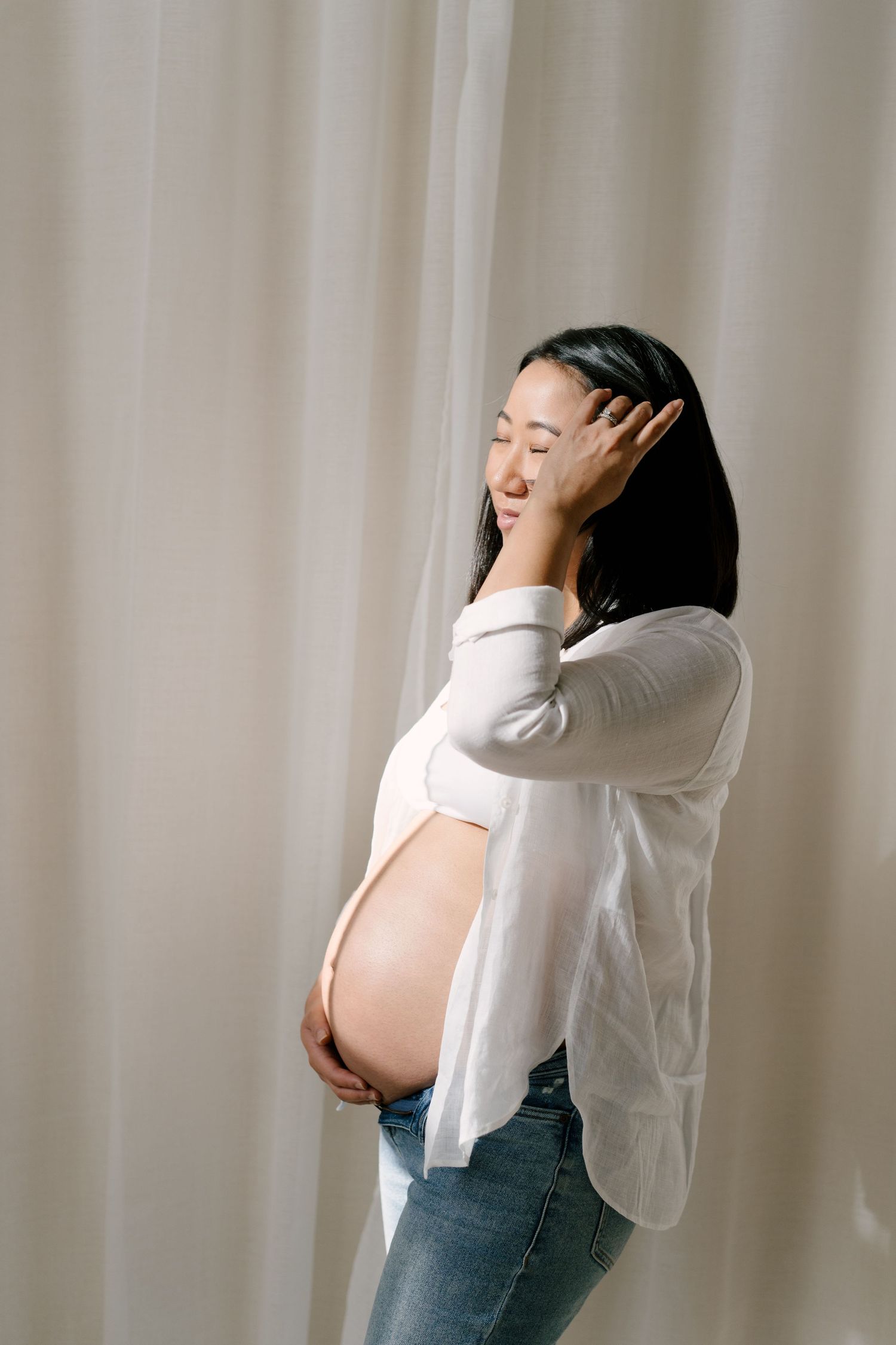 Pregnant woman in white shirt cradles baby bump with eyes closed, standing by soft white curtains.