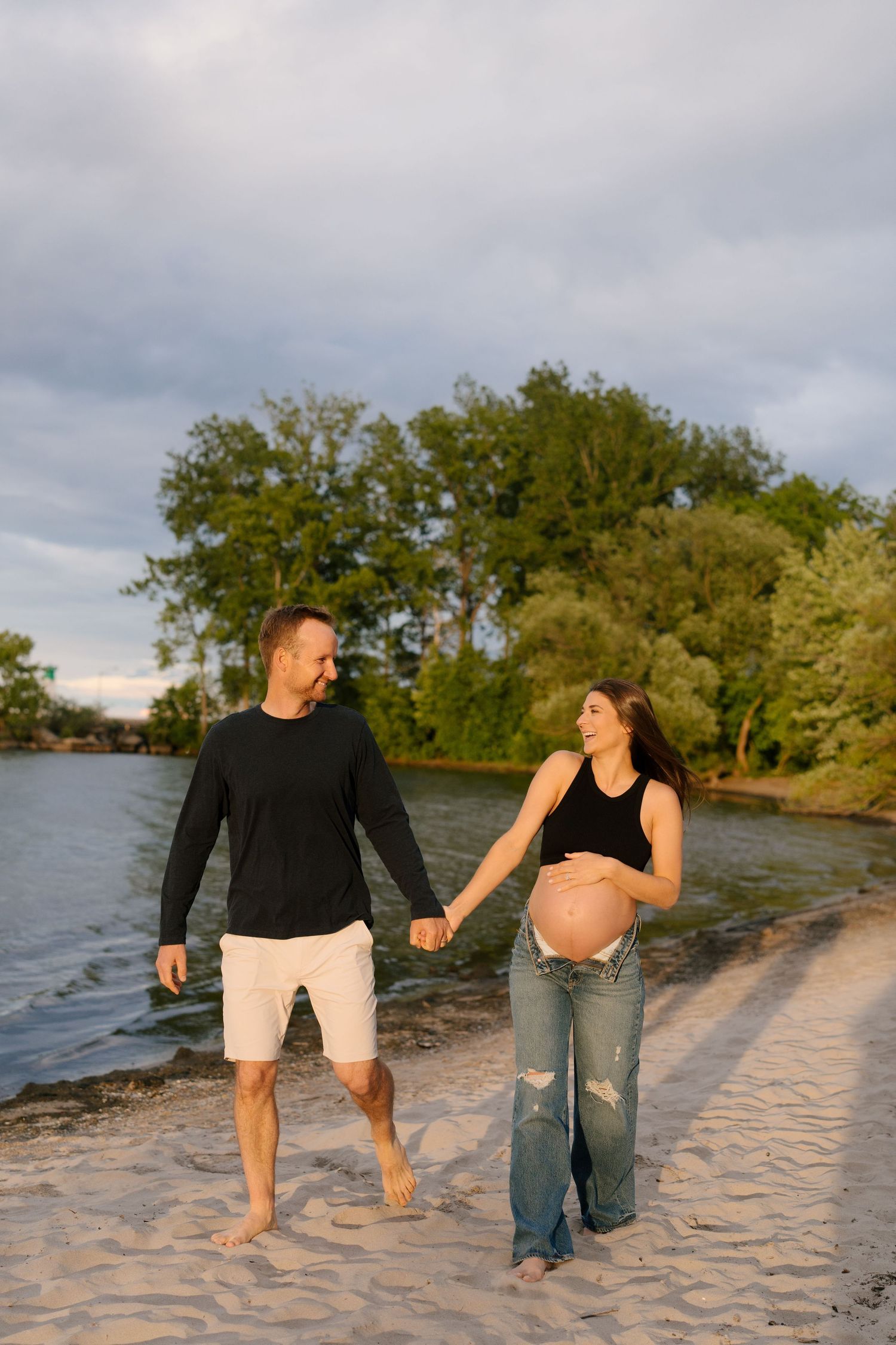 Smiling pregnant couple walks hand-in-hand along sandy beach at sunset, woman holding her baby bump tenderly.