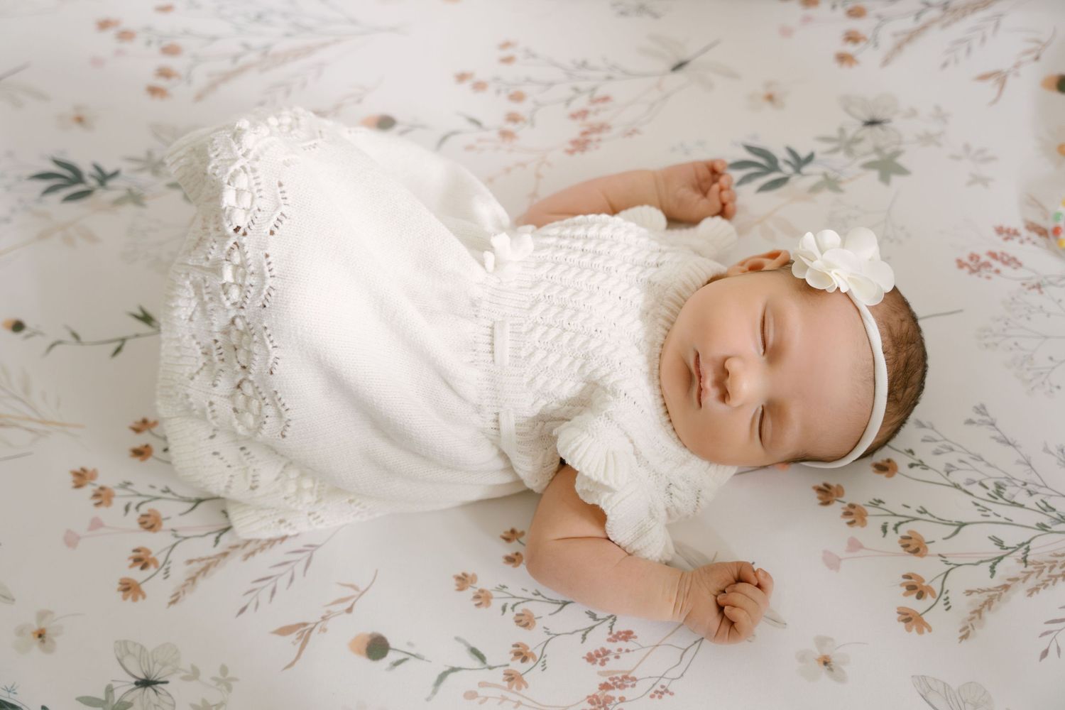 Sleeping newborn baby girl in white knit dress with floral headband lying on floral blanket.