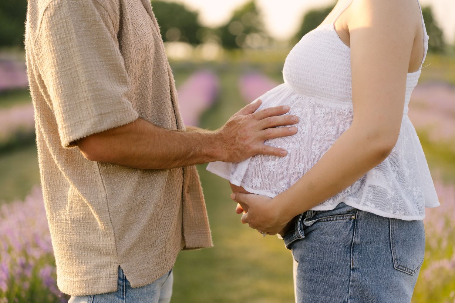 Close-up of couple gently cradling pregnant belly together in a purple lavender field at sunset.