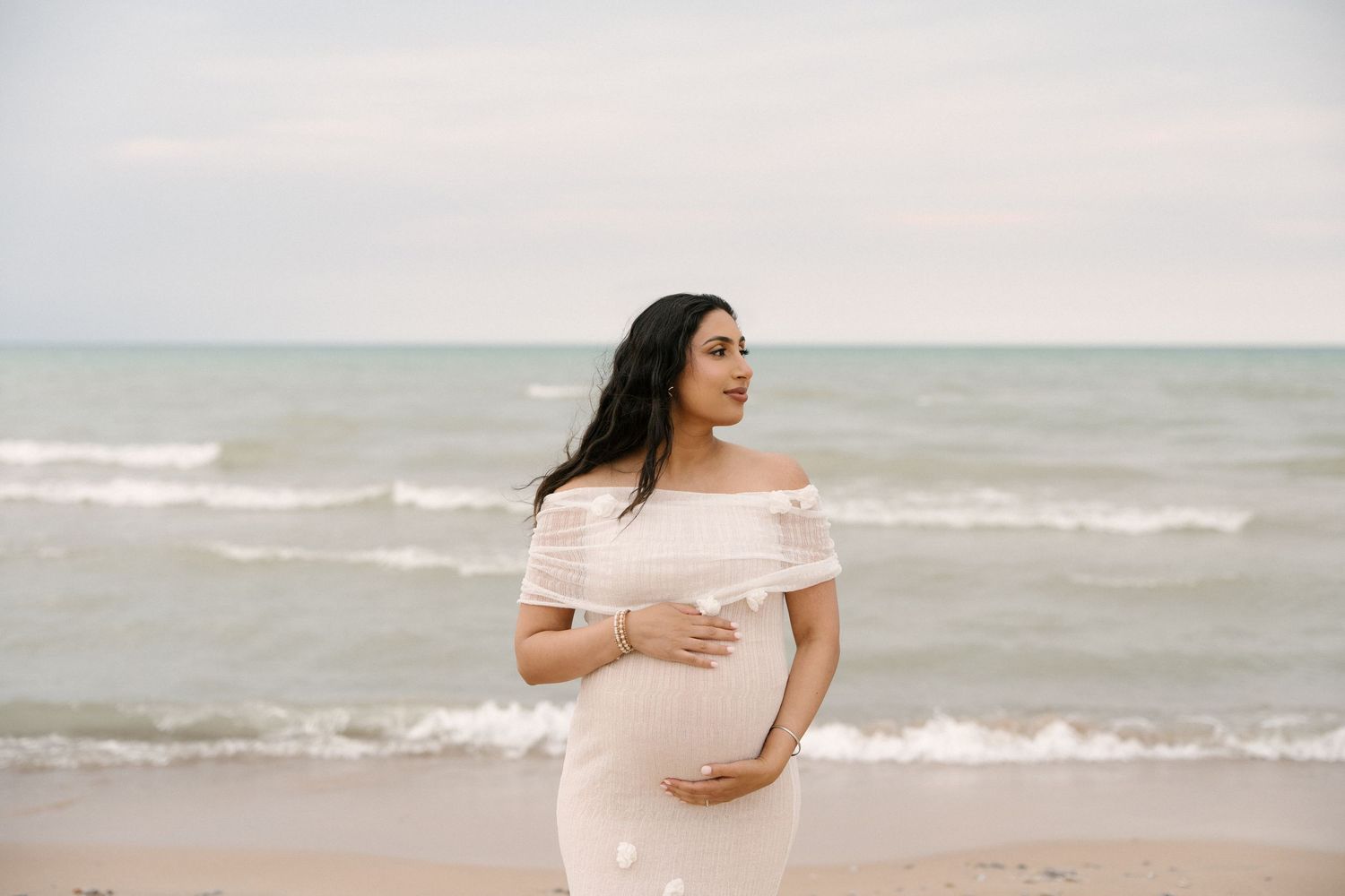 Pregnant woman in white lace off-shoulder dress cradling baby bump on serene ocean beach.