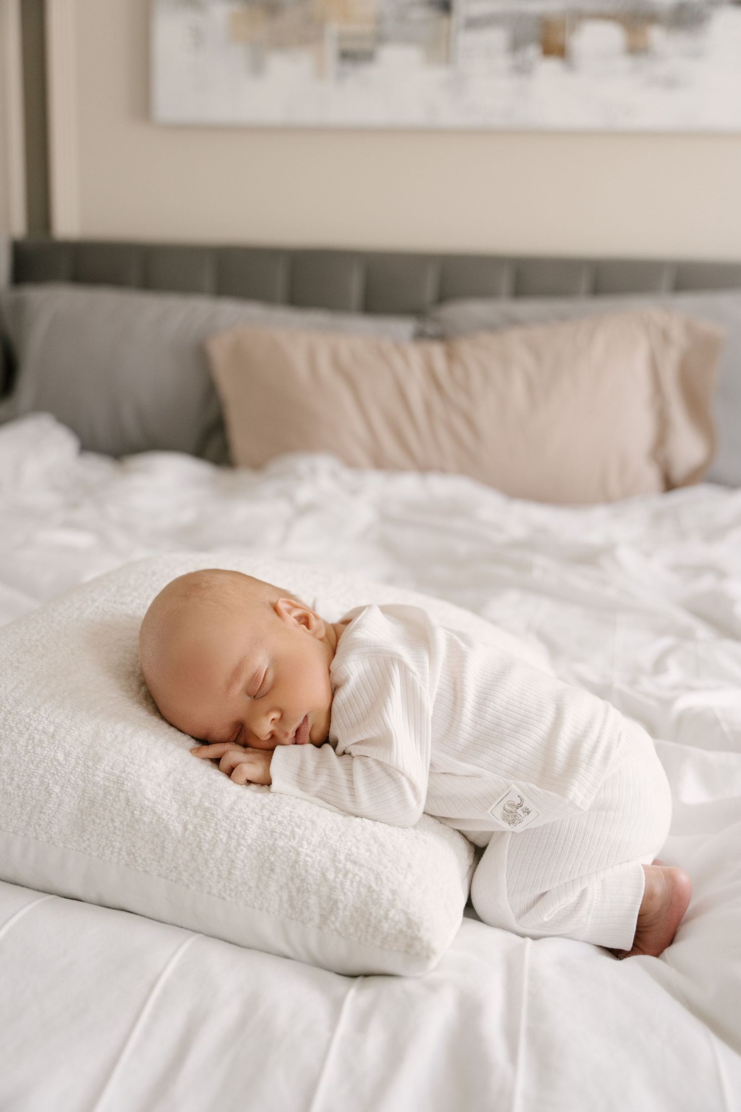 A peaceful newborn baby in white onesie sleeps curled up on a soft pillow on a white bed.