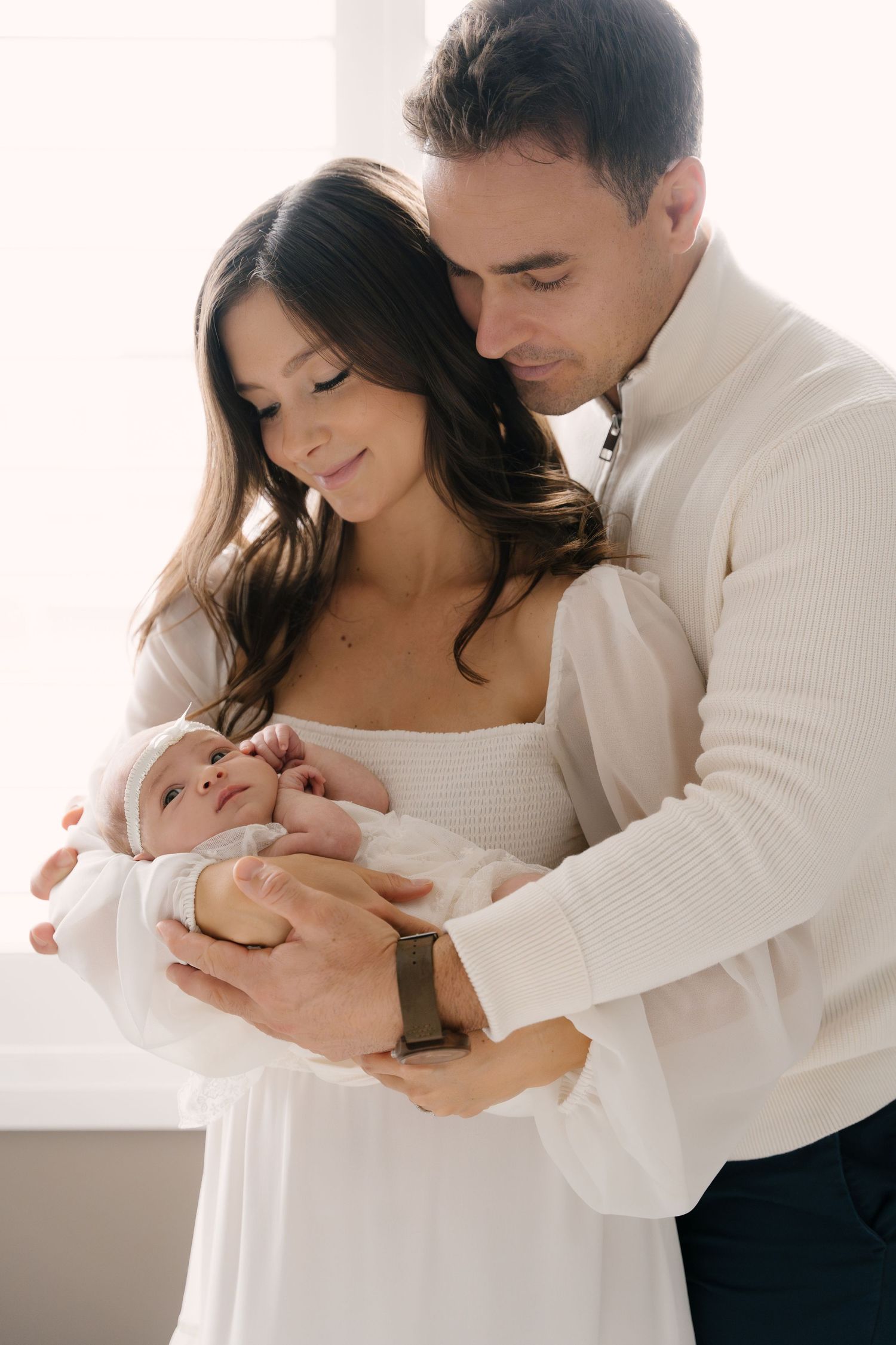 Young couple in white outfits tenderly holding their newborn baby indoors.