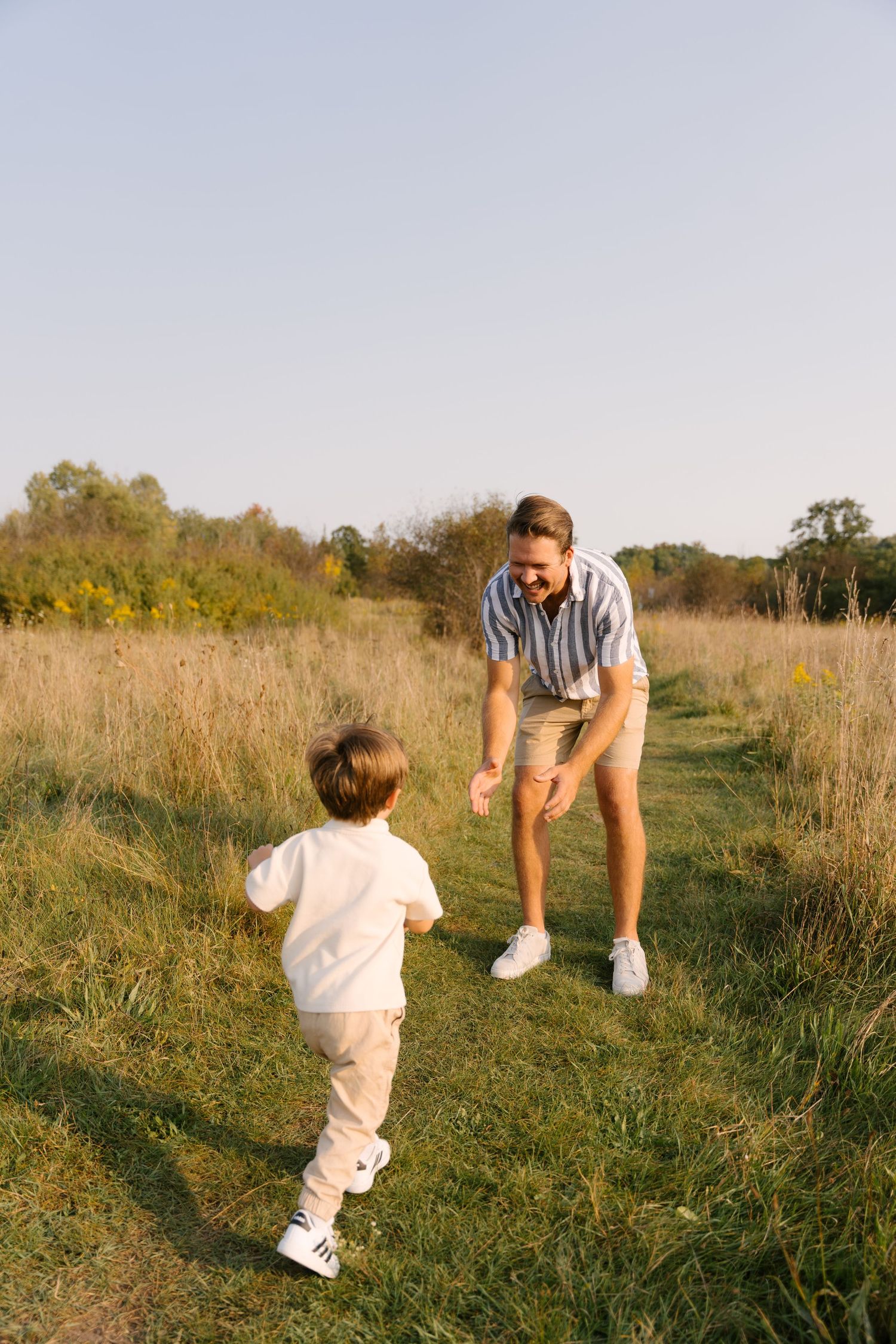 Father bends down to greet his young son running toward him on a grassy field path at golden hour.