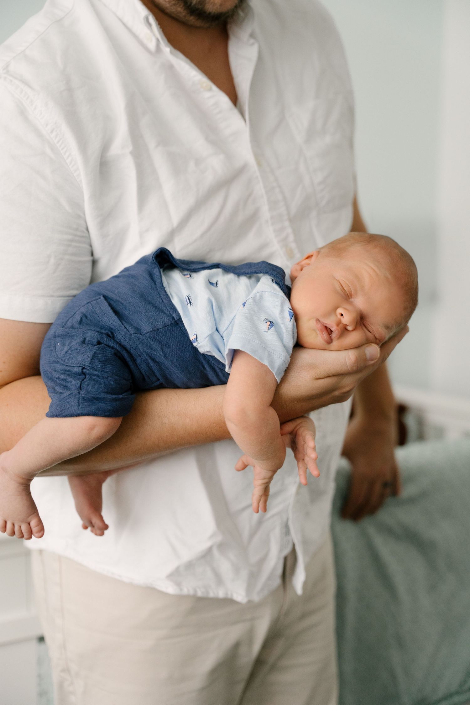 Father in white shirt gently cradles sleeping newborn baby in his arms.