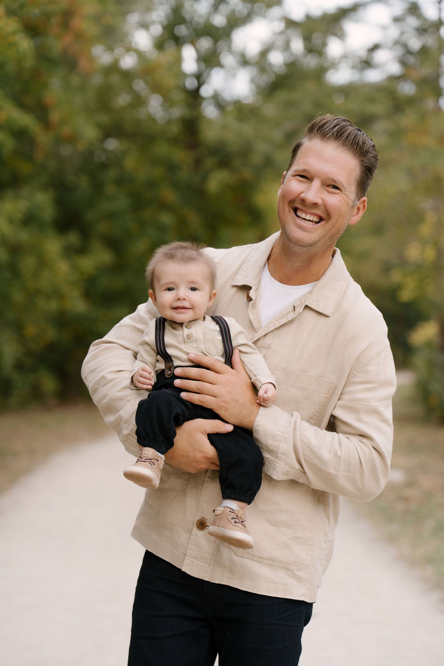Cheerful father in beige shirt holds his baby boy while smiling on a tree-lined outdoor path.