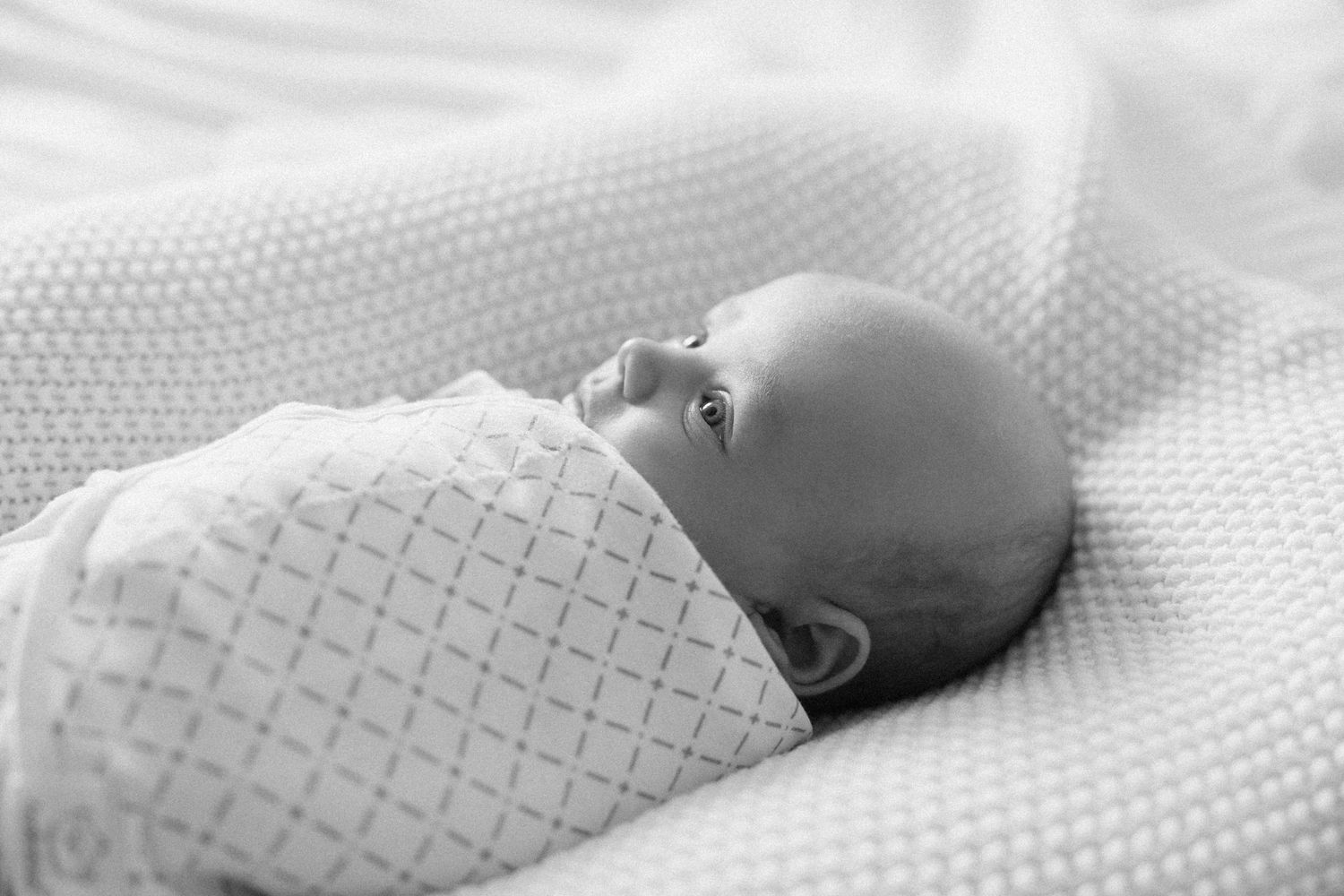 Newborn baby swaddled in white blanket lying peacefully on soft textured surface, black and white photo.