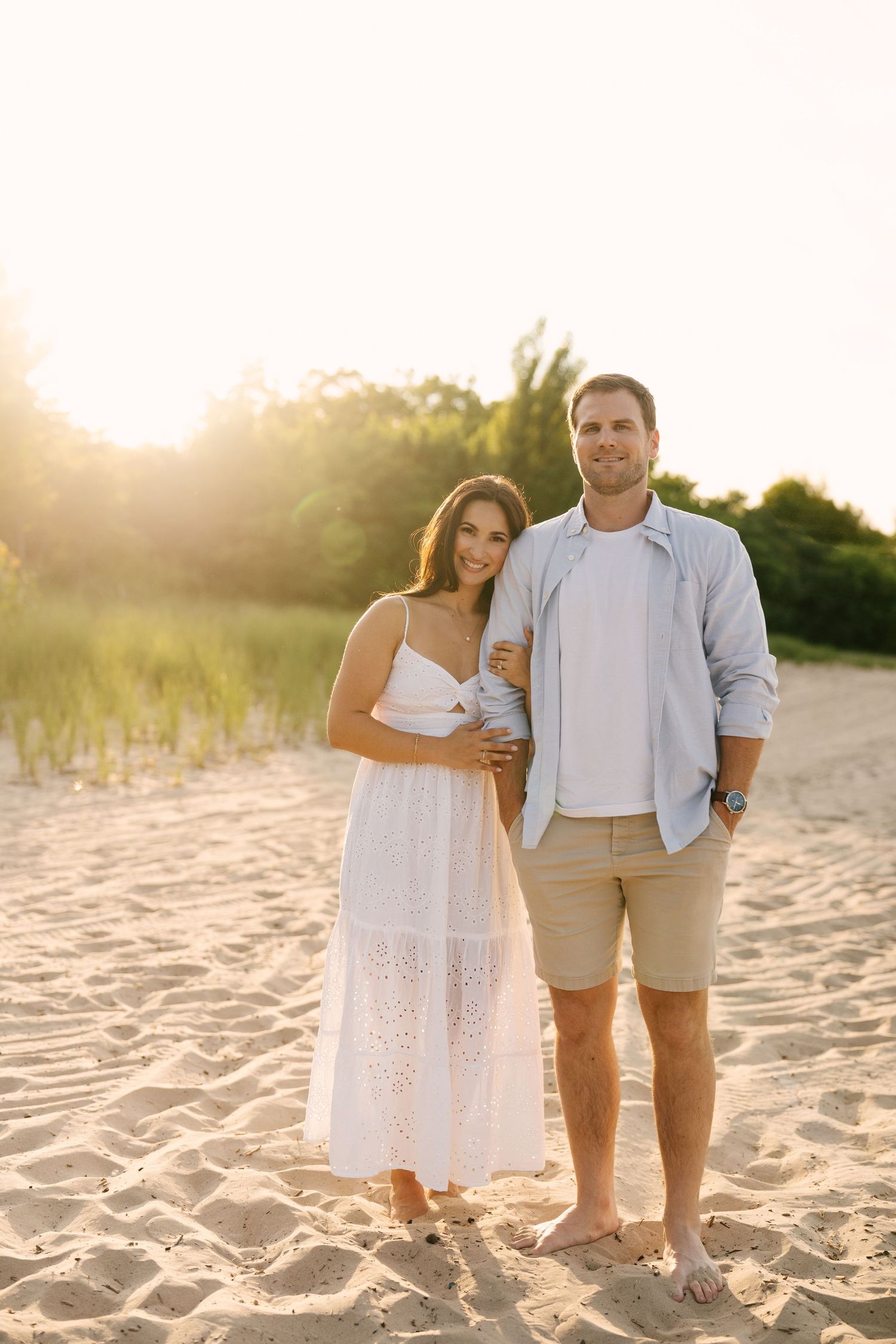 Smiling couple posing together on sandy beach at golden sunset, woman in white dress, man in light blue shirt.