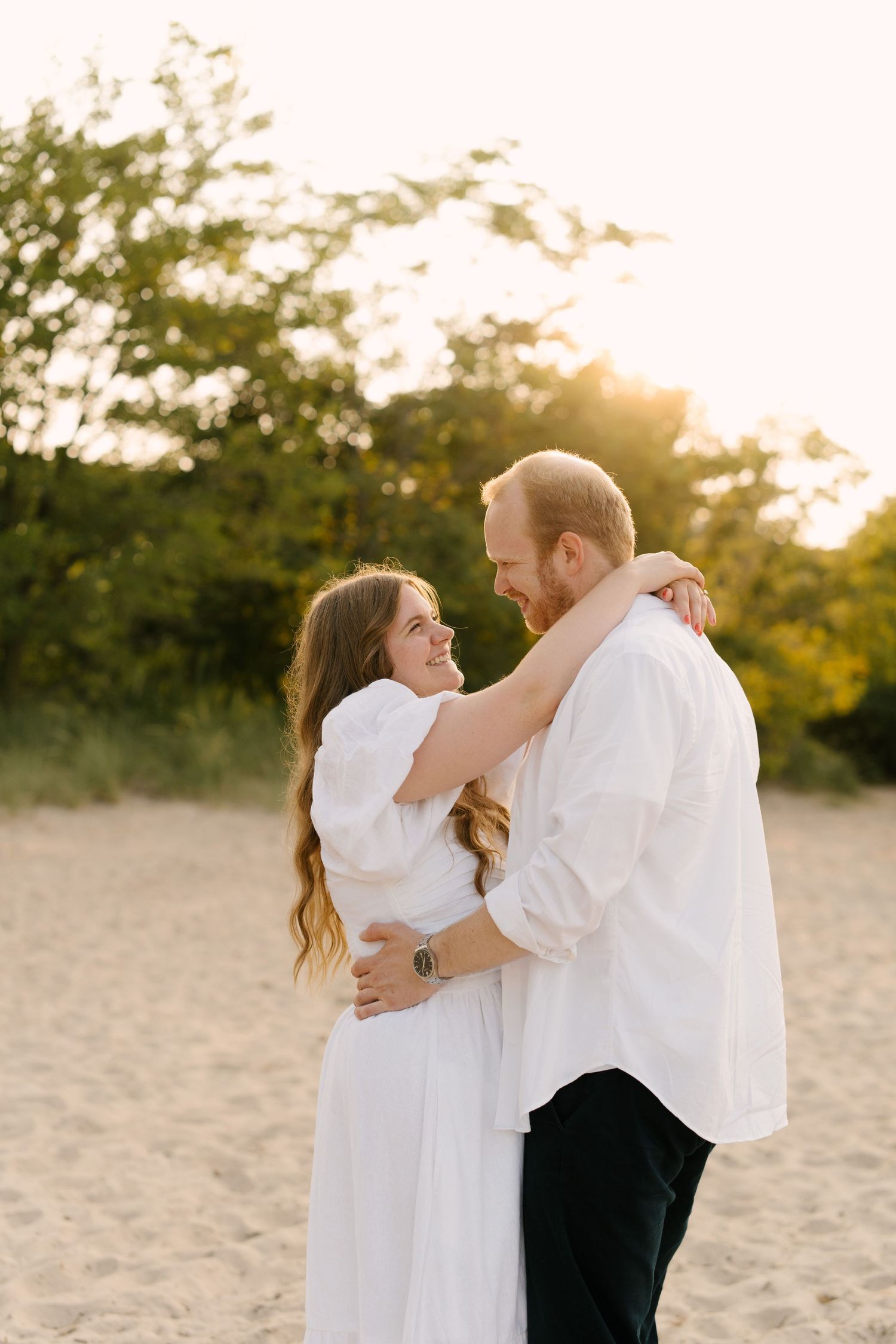 Couple embracing on sandy beach at golden sunset, woman in white dress smiling up at bearded man in white shirt.