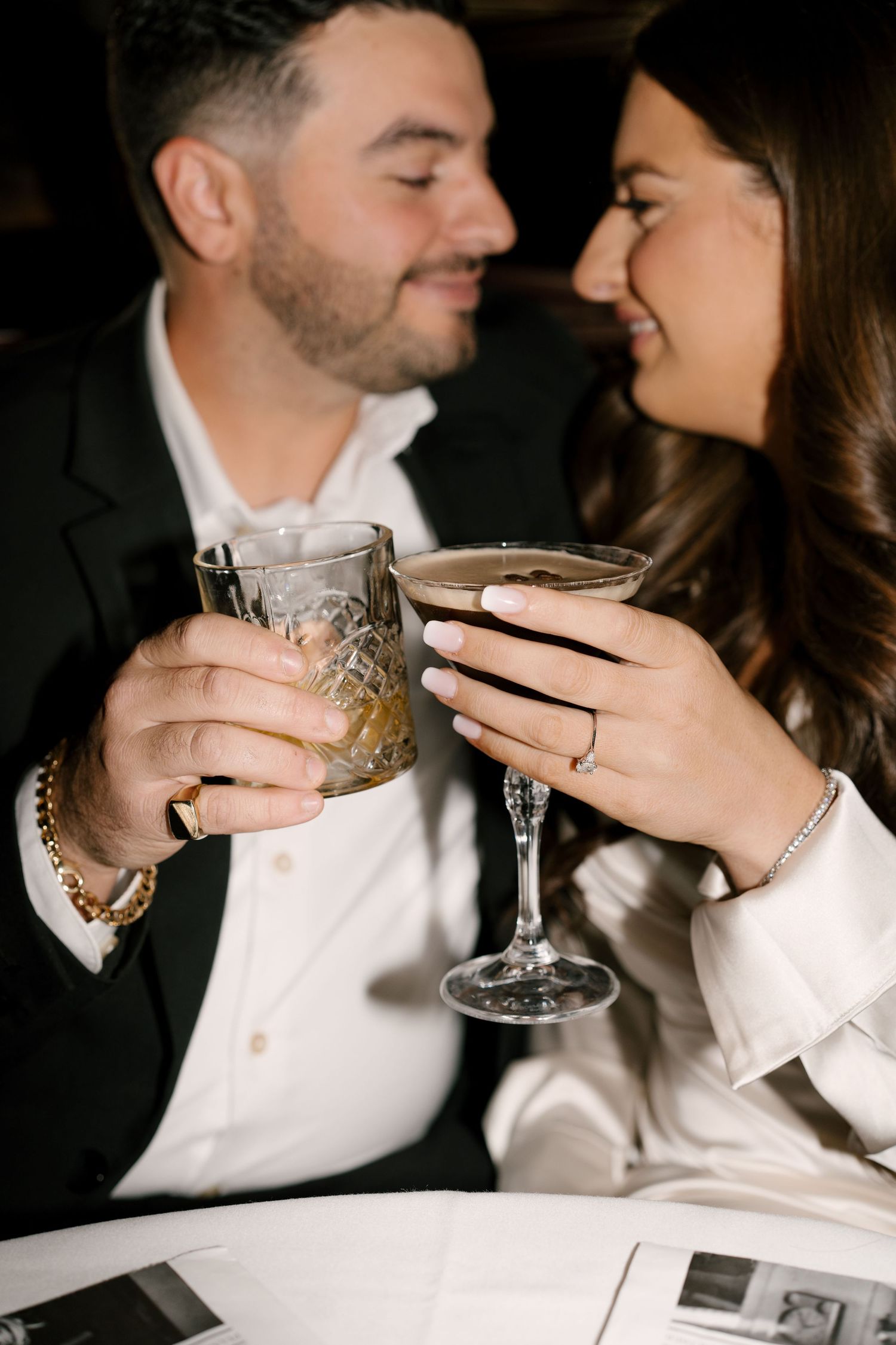 Engaged couple clinking cocktail glasses together intimately at a romantic dimly lit restaurant.