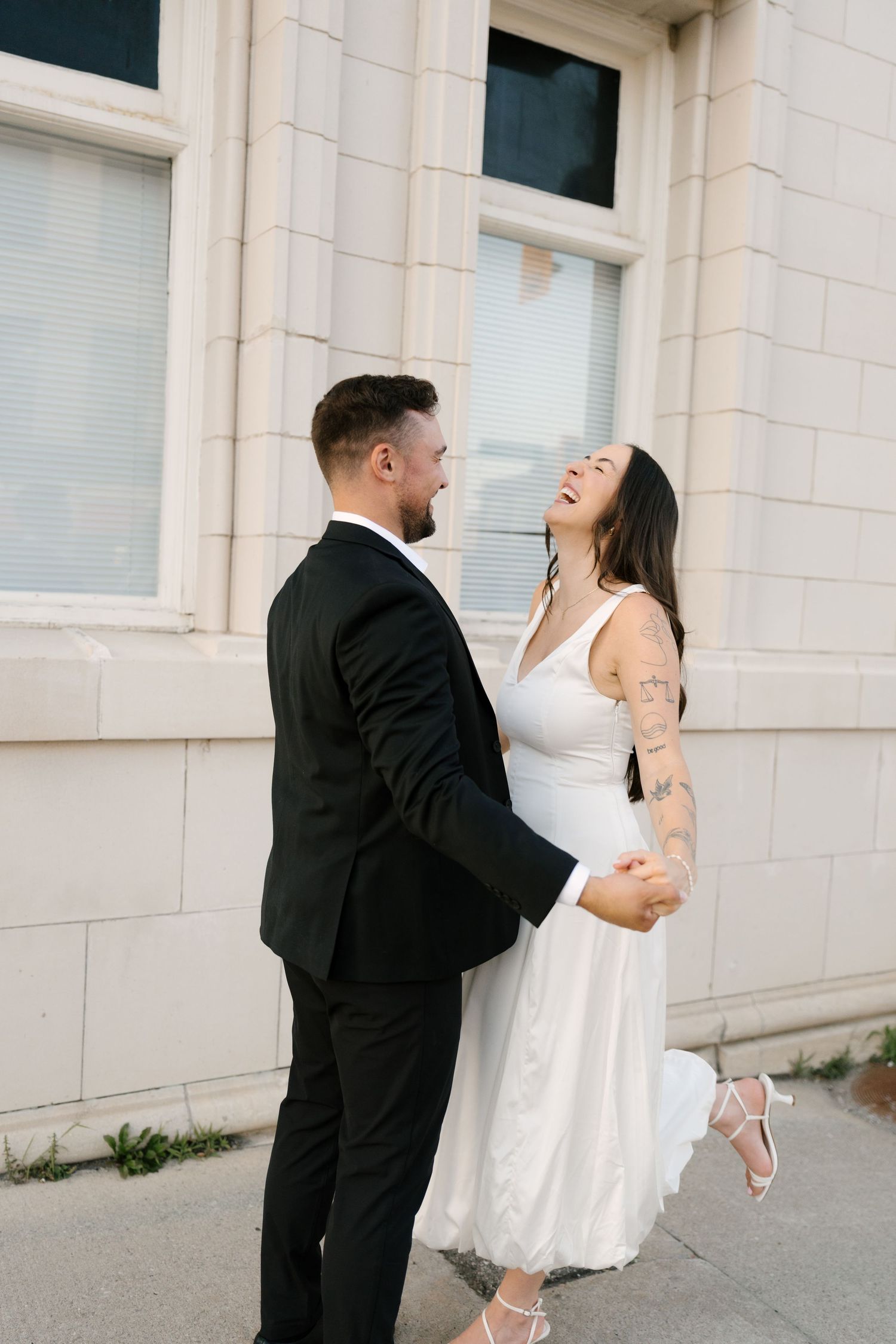 Bride laughing joyfully while holding groom's hands outside a classic stone building.