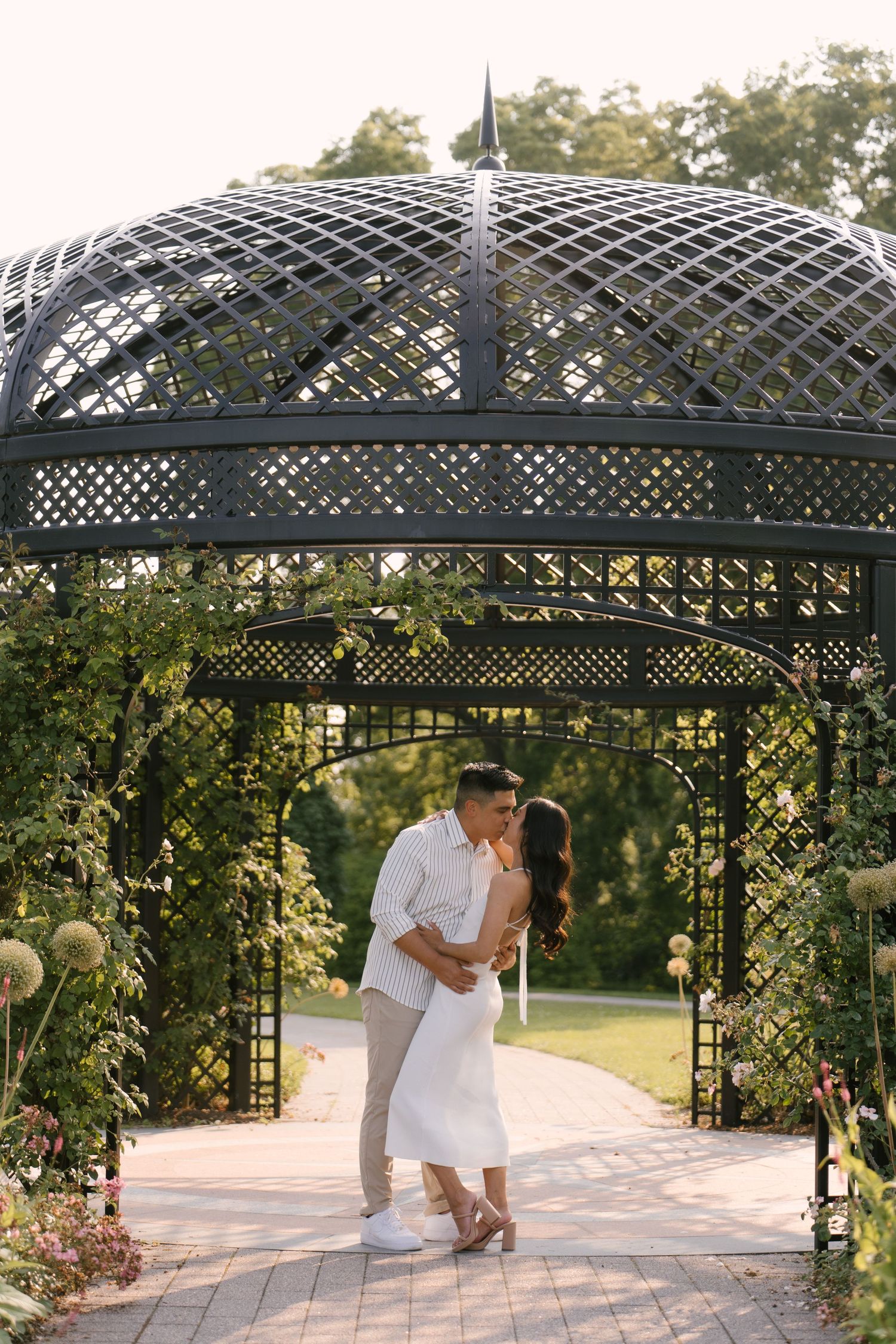 Romantic couple sharing a kiss under an ornate black lattice garden gazebo surrounded by flowers.