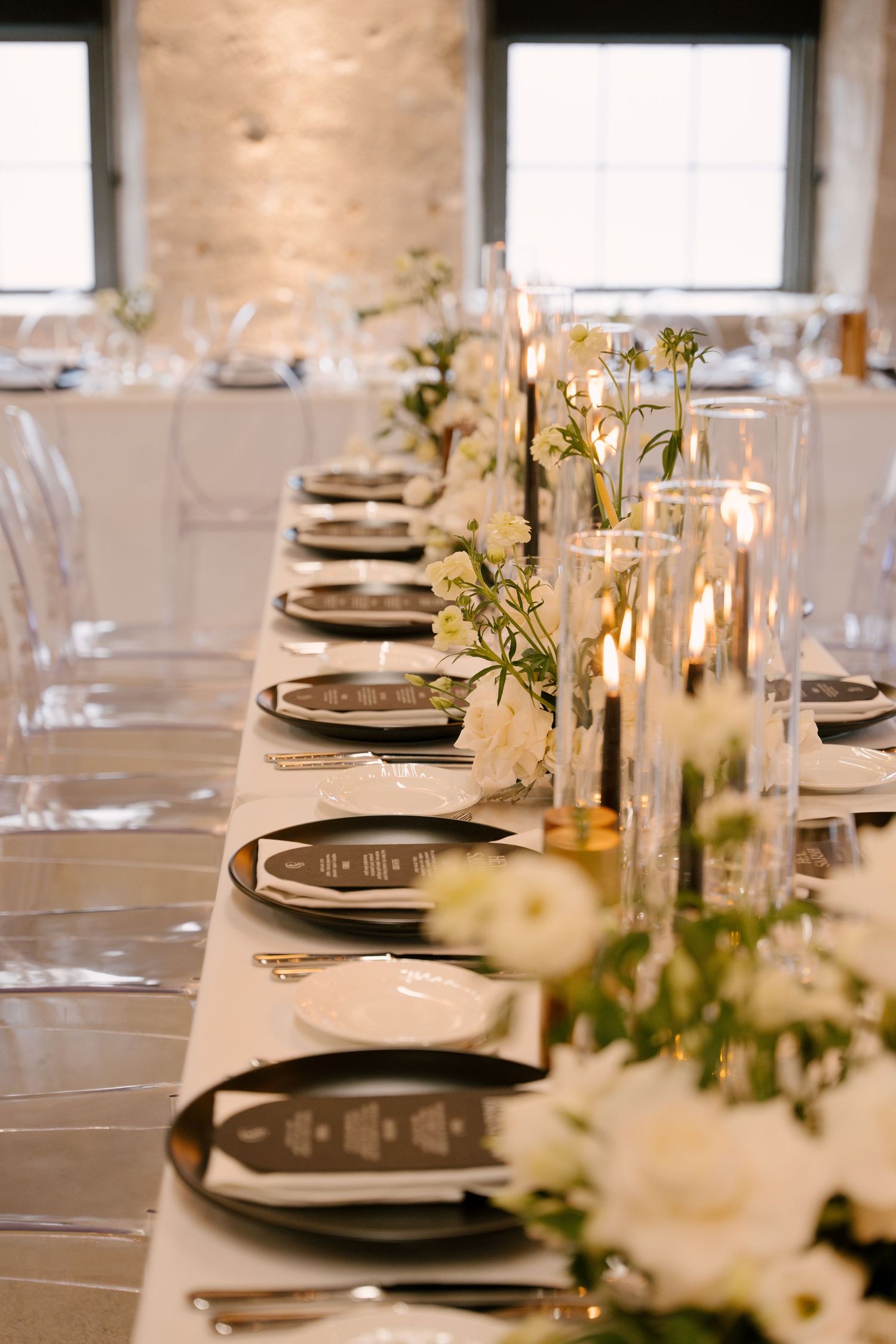 Long wedding reception table with black charger plates, white florals, and tall glass candle holders at Tapestry Hall.