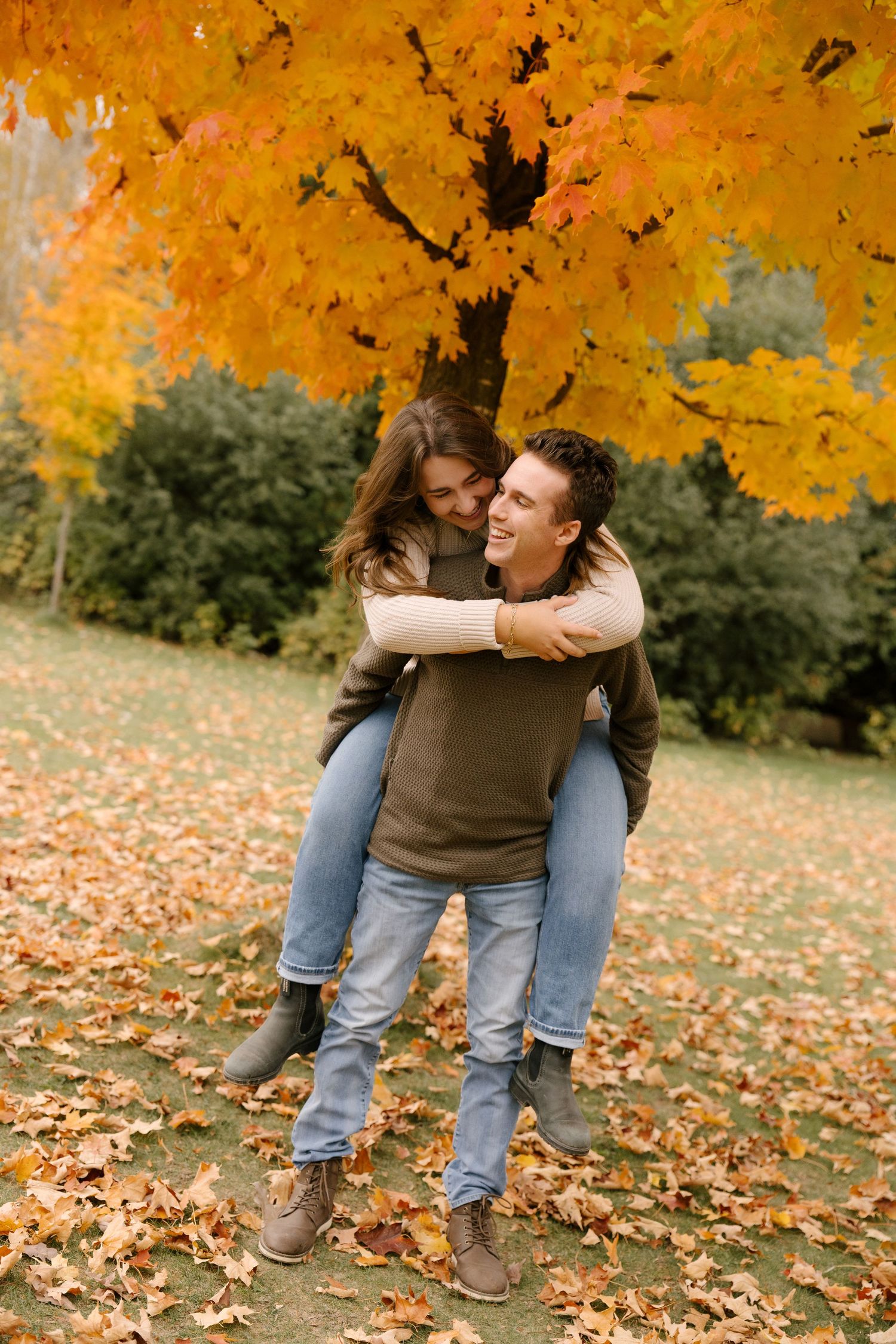Joyful couple laughing outdoors in autumn park with man giving woman a piggyback ride.