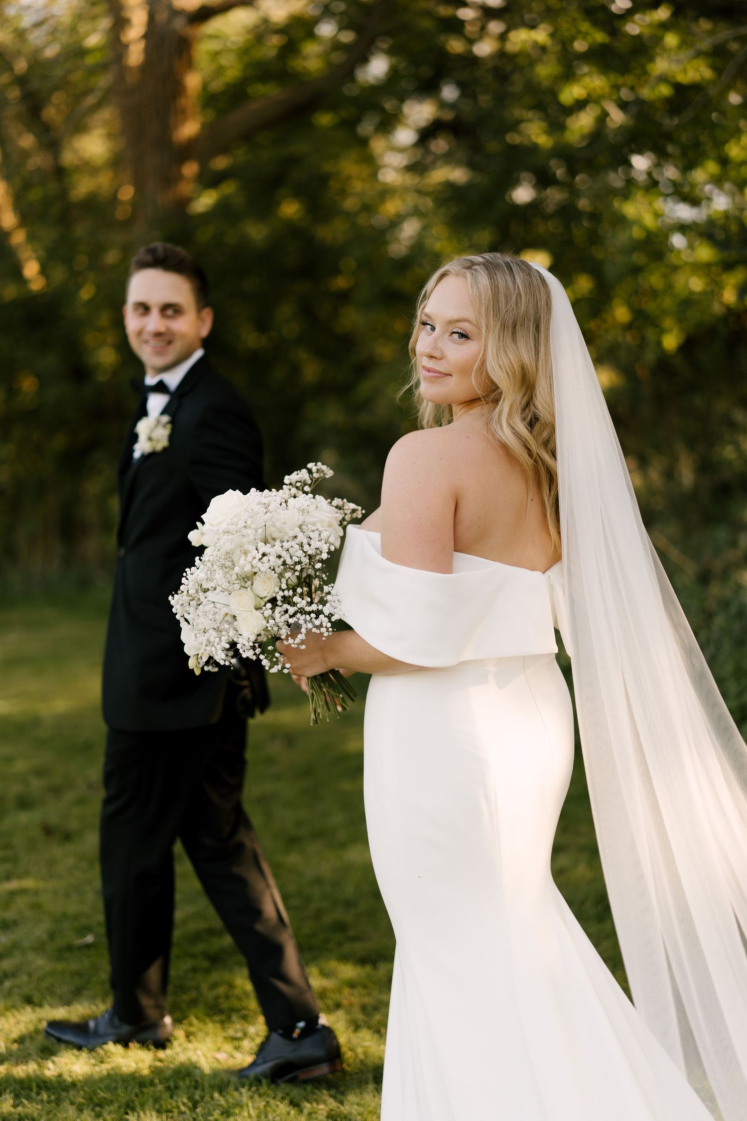 Bride in fitted white gown and veil glances back while groom stands in black tuxedo on lush green lawn.