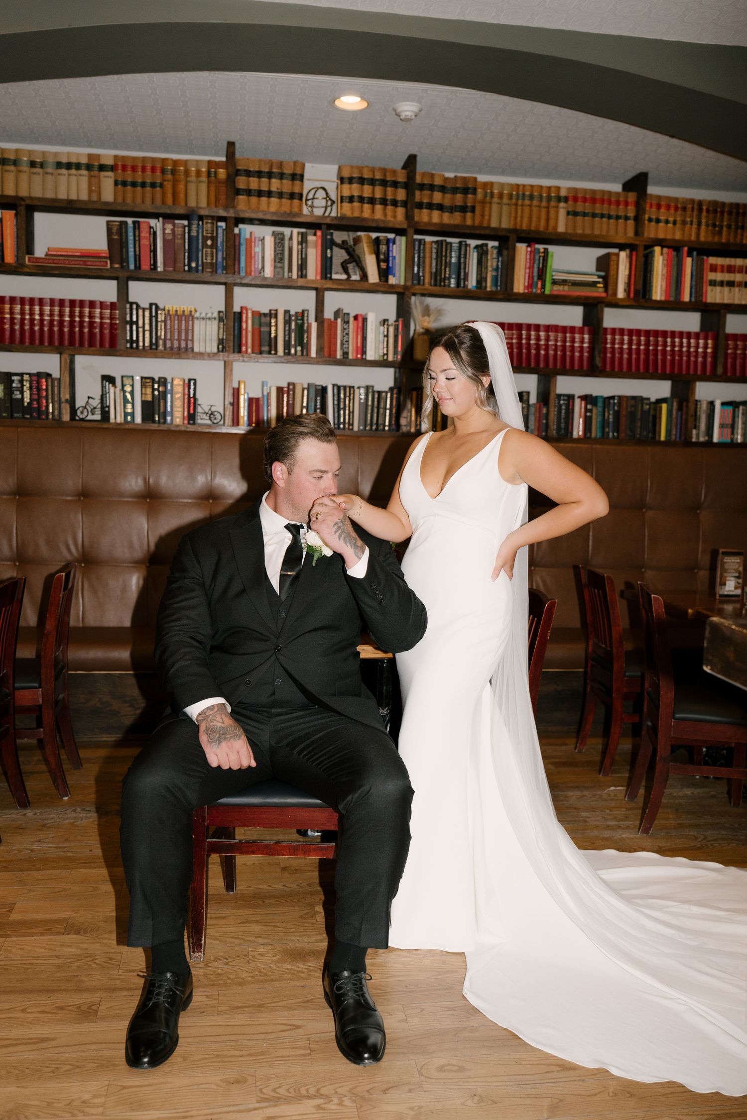 Groom kisses bride's hand in library setting with bookshelves in background.