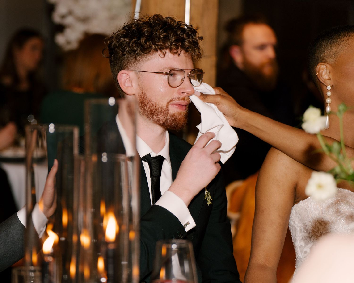 Emotional groom wiping tears at wedding reception while seated at elegantly decorated dinner table with candles.